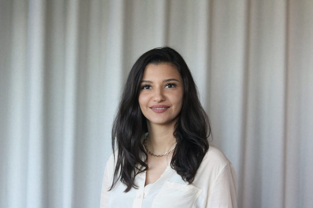 Eine lächelnde Frau mit schulterlangem dunklem Haar und weißem Hemd steht vor einem hellgrauen Vorhang in einem Coworking Space. Sie wirkt selbstbewusst und entspannt und blickt direkt in die Kamera. An image from EDGE Workspaces.