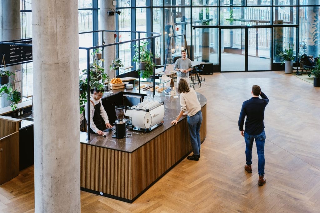 Ein geräumiges, modernes Café mit großen Fenstern und Grünpflanzen dient auch als Coworking Space. Vier Personen arbeiten um eine zentrale Kaffeebar mit Holzverkleidung herum: ein Barista bereitet den Kaffee zu, während zwei Ein Bild von EDGE Workspaces.