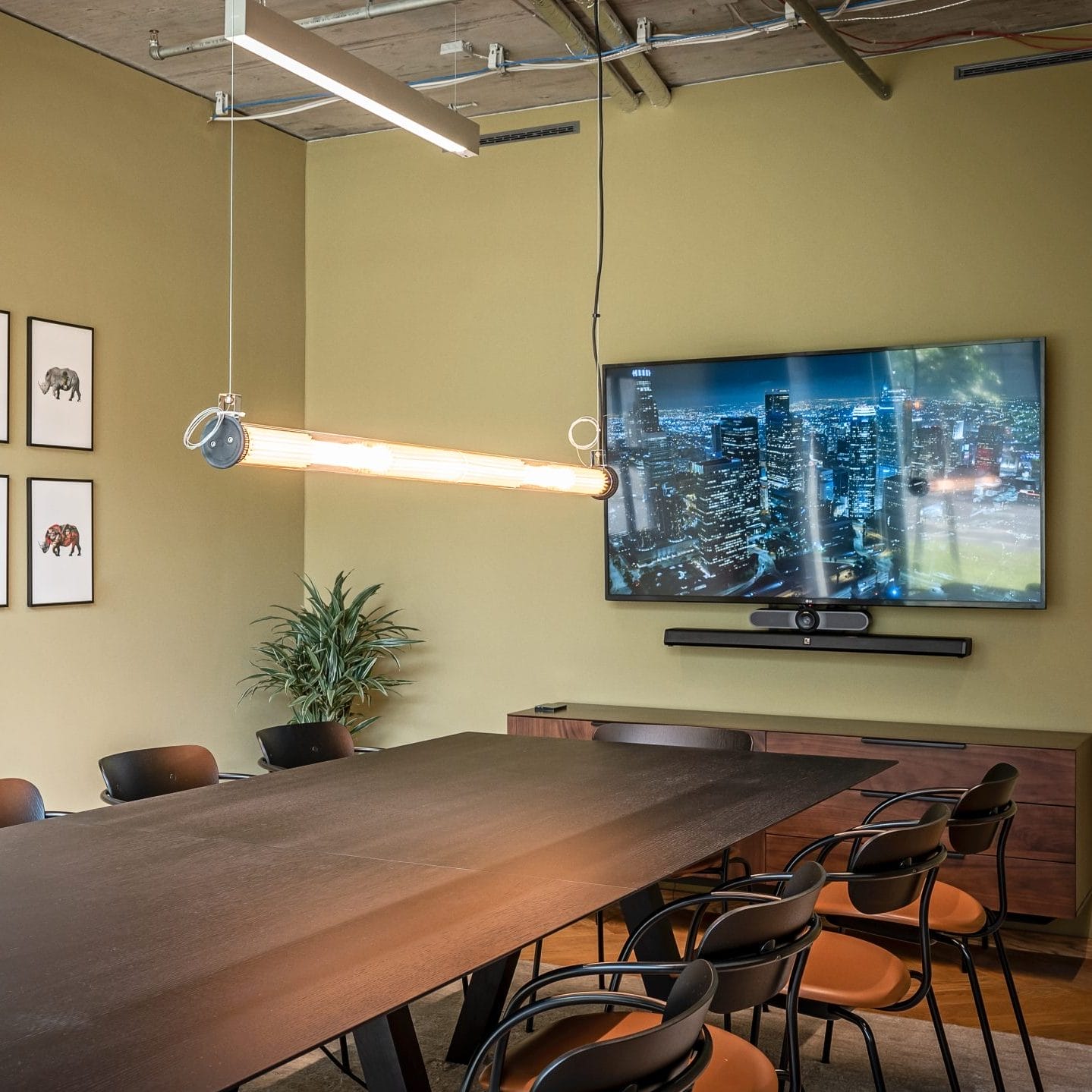 A modern conference room featuring a long, dark wooden table surrounded by black chairs. Walls are olive green, and decorative art hangs on one side. A large TV displays a cityscape. Overhead, An image from EDGE Workspaces.