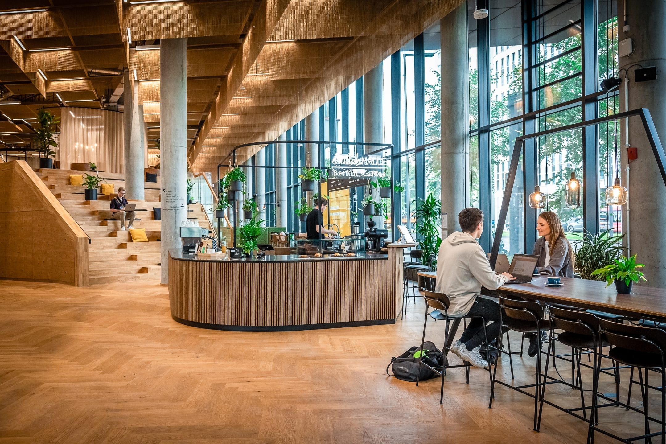 Modern office café with wooden interiors, large windows, and terrarium-style pendant lights. This flexible workspace solution allows people to be casually seated, some working on laptops. A curved barista counter is visible An image from EDGE Workspaces.