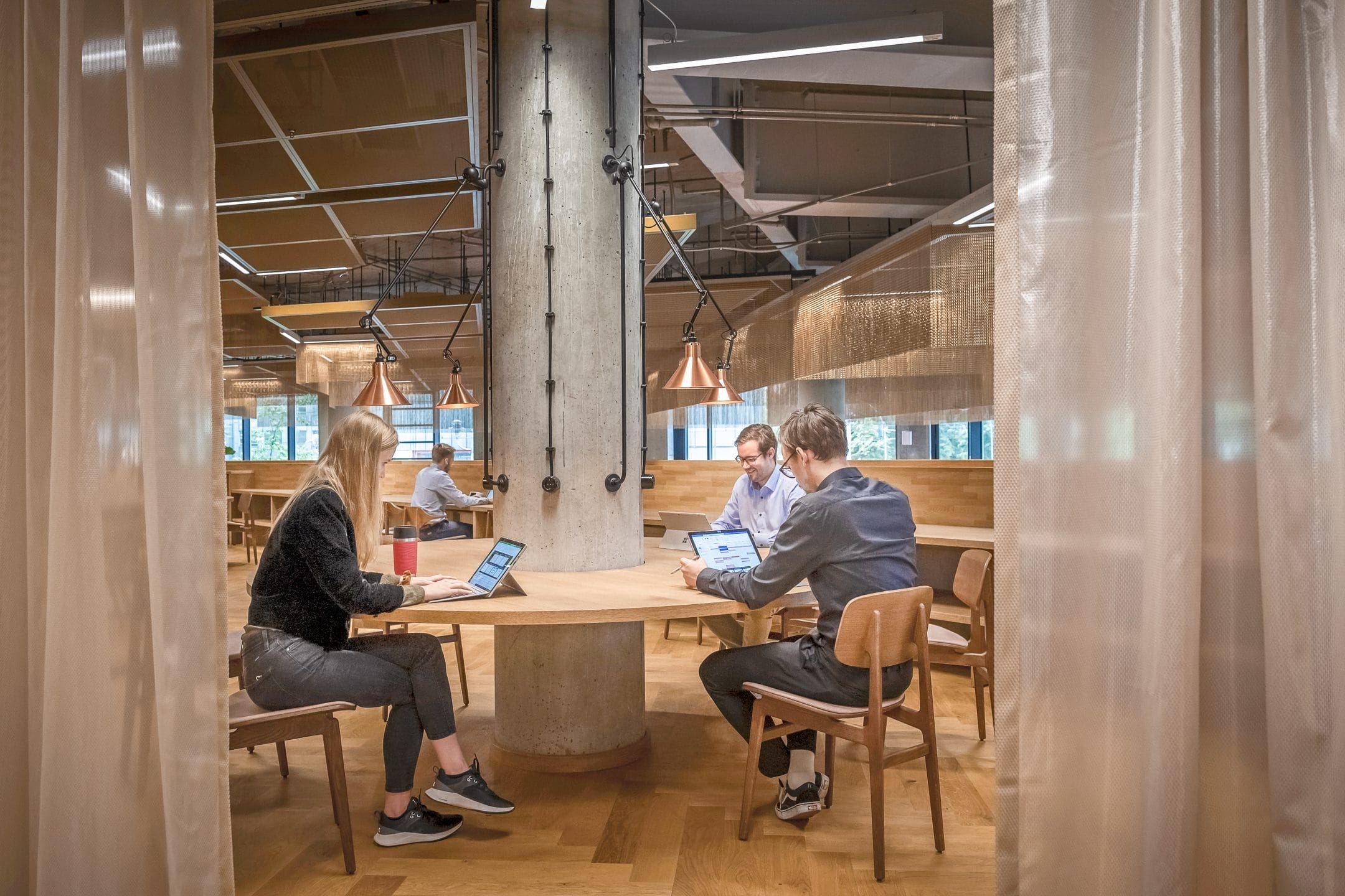 Three professionals, two men and one woman, sit at a wooden table in an innovative coworking environment, using laptops and discussing work. The area features exposed beams, stylish lighting, and semi-transparent dr An image from EDGE Workspaces.