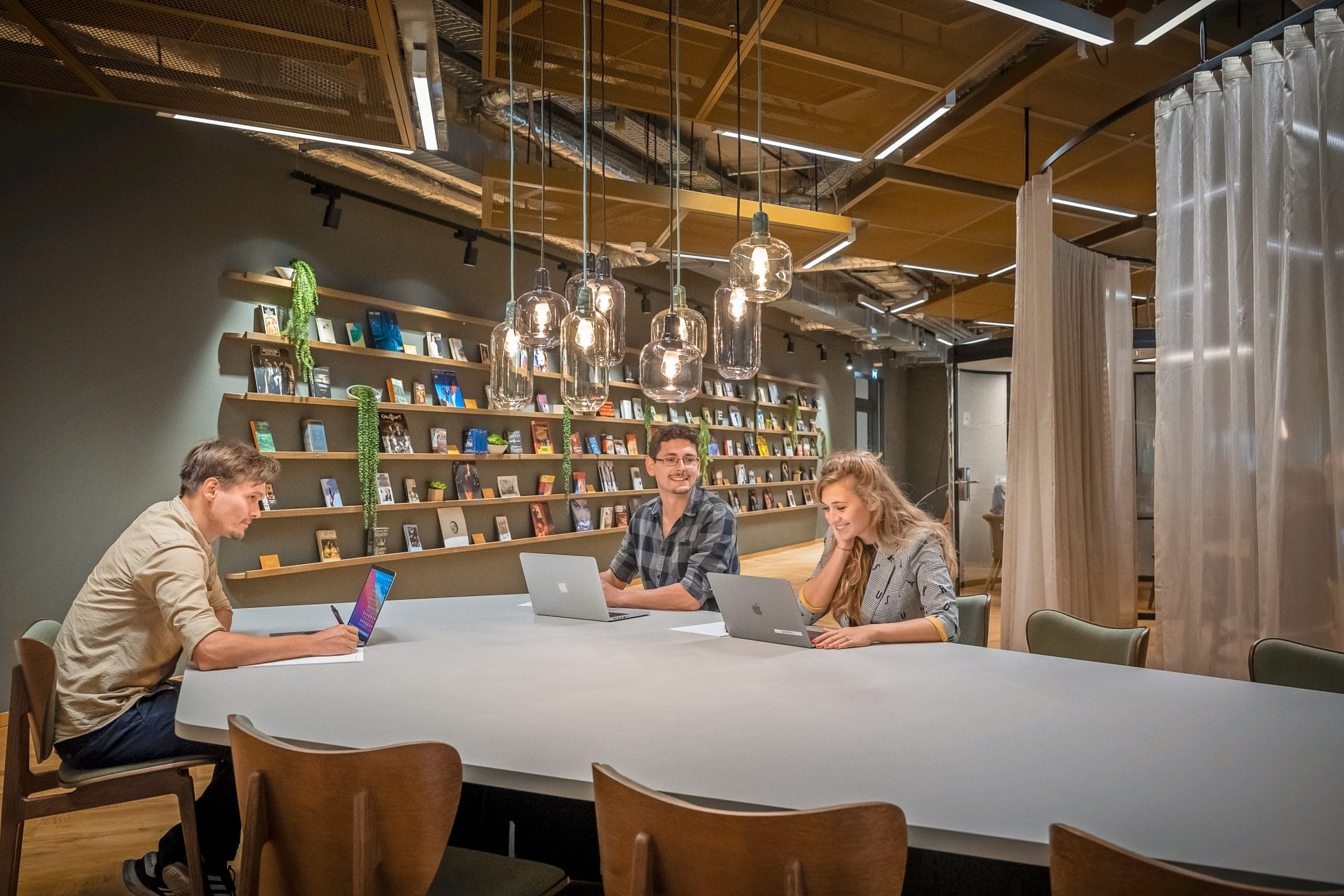 Three people, two men and one woman, are seated at a long white table in a modern office space, working on laptops and a tablet. The room features stylish hanging lights and a bookshelf filled An image from EDGE Workspaces.