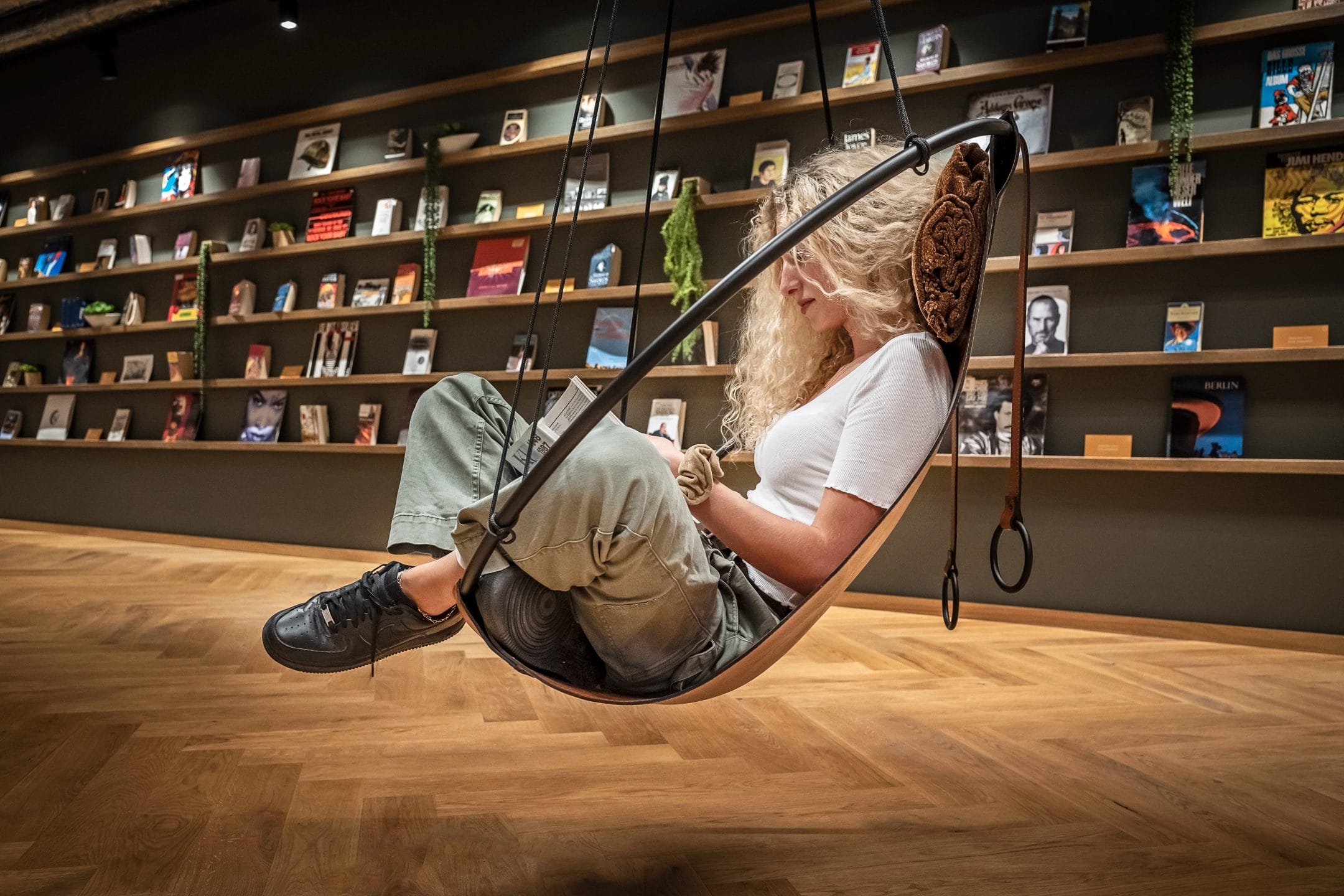 A woman sits in a stylish swing chair, reading a book in a modern bookstore that doubles as an innovative coworking environment, with walls lined by shelves filled with books. Her relaxed pose and focus denote An image from EDGE Workspaces.