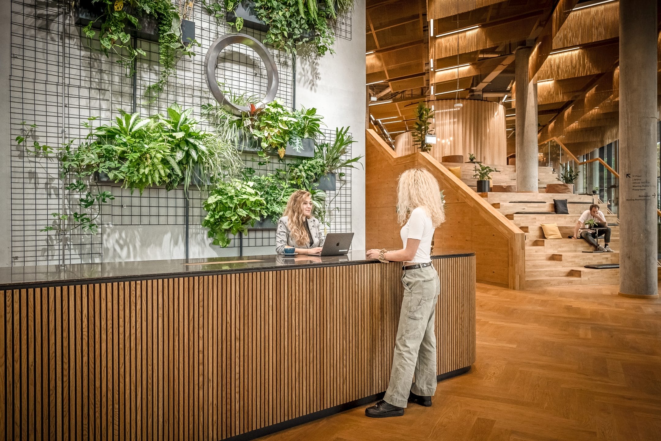 Two women converse at a modern reception desk in a spacious office foyer with wooden stairs, abundant green plants, and relaxed seating areas in the background, showcasing sustainable office solutions. An image from EDGE Workspaces.