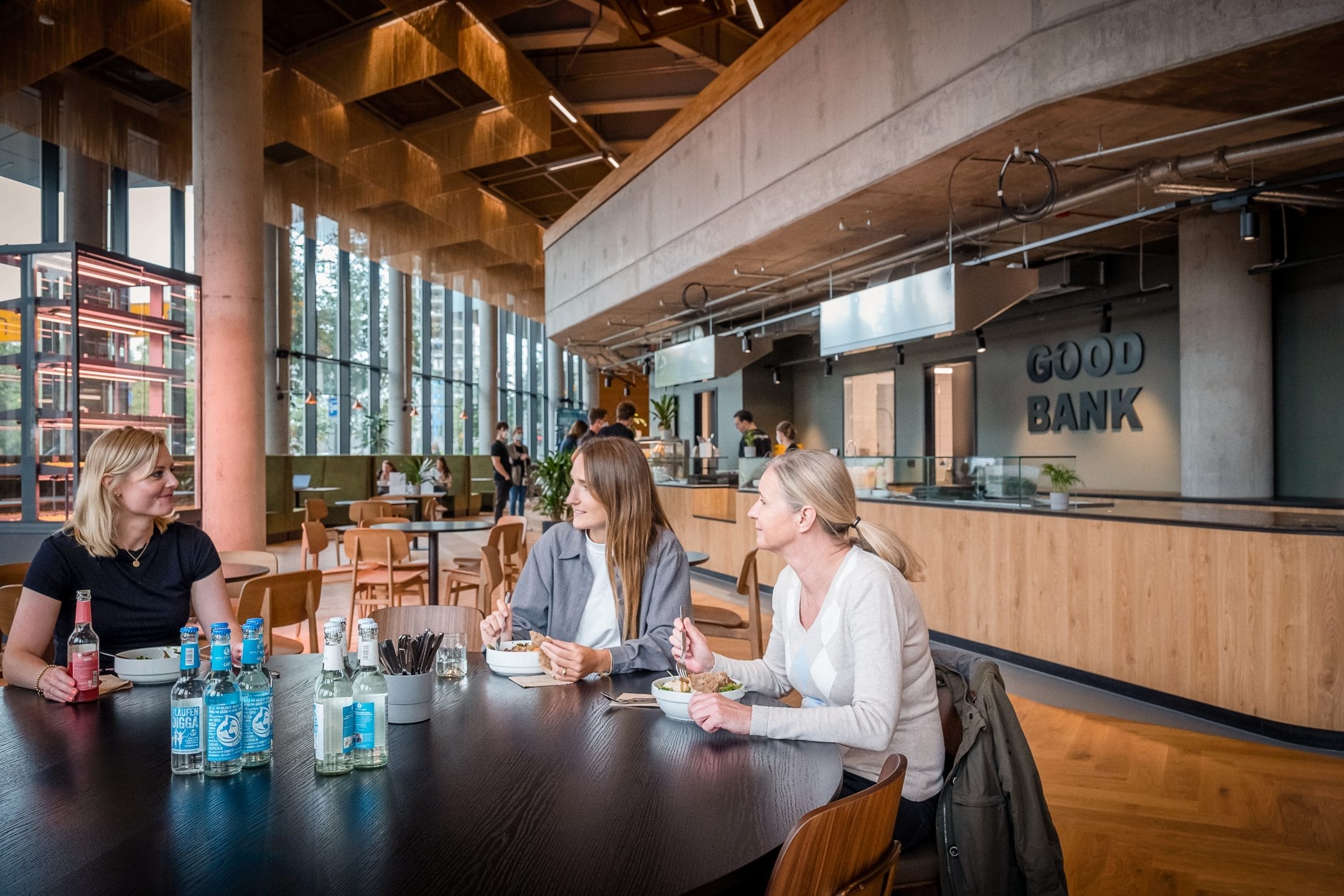 Three women sit at a table in a modern café with "good bank" written on the wall behind them. They are conversing and holding beverages, surrounded by other patrons and a spacious wood-themed interior An image from EDGE Workspaces.