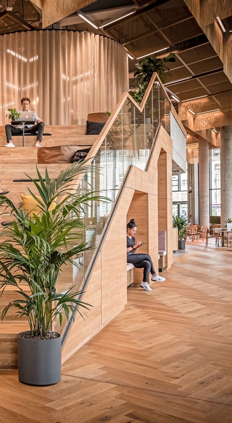 Ein moderner Büroraum, ausgestattet mit großzügigen Holzböden und Treppen, ist auf Flexibilität ausgelegt und bietet die Möglichkeit für private Büros und Mitgliedschaften. Zwei Personen benutzen Laptops; eine sitzt auf der Treppe und eine andere An image from EDGE Workspaces.
