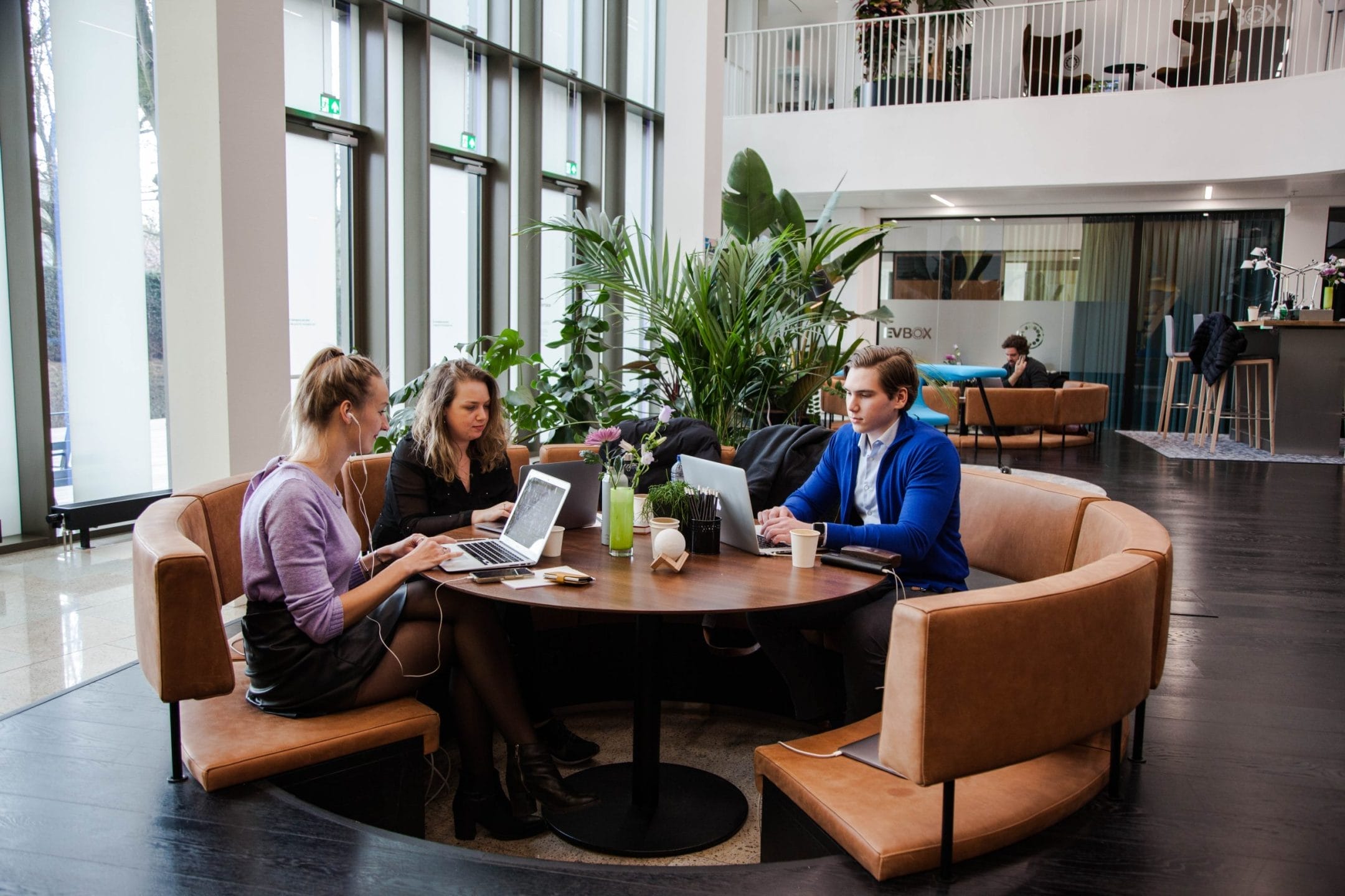 Three professionals, two women and one man, sit around a circular table in an innovative coworking environment, working on laptops and discussing. Large windows and green plants are visible in the background. An image from EDGE Workspaces.