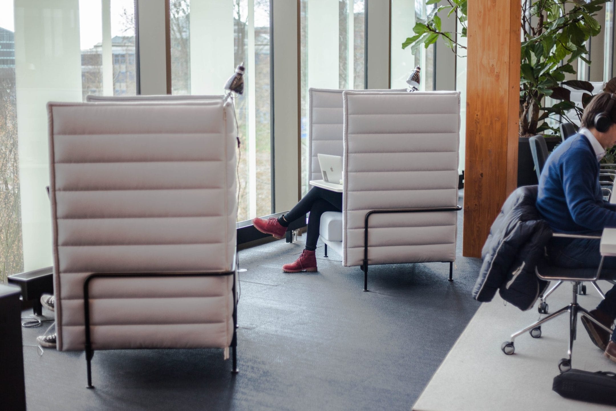 A modern office setting with two high-back chairs in the foreground. A person is visible between the chairs, sitting near a window, while another works at a desk with headphones. The room has ample natural An image from EDGE Workspaces.