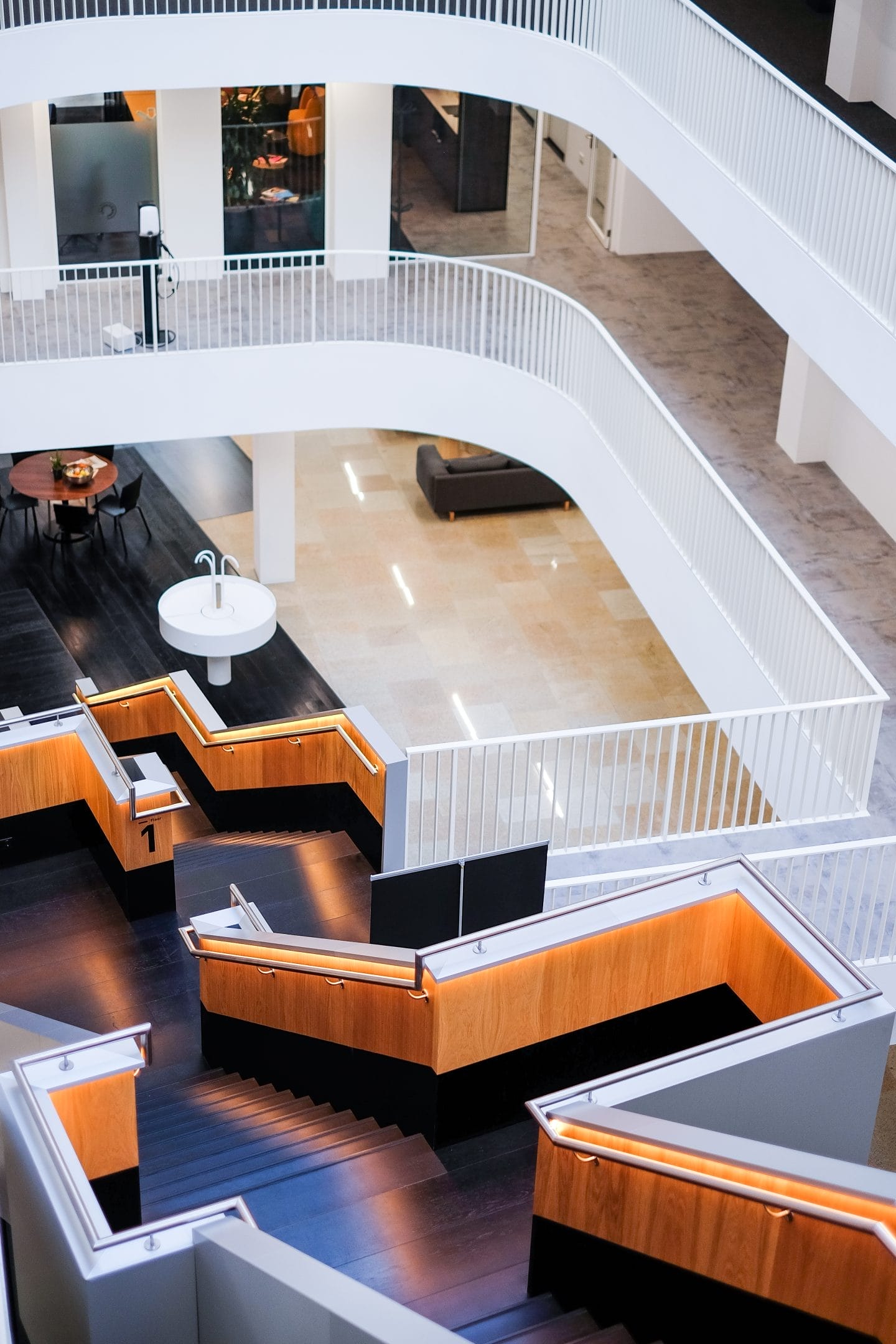 A modern office building interior featuring multiple levels with sleek white staircases and wooden accents. the space includes various workstations with black chairs and desktop lamps, set in an open and airy coworking layout. An image from EDGE Workspaces.