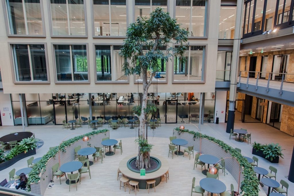 Interior view of a modern office atrium with large potted trees, surrounded by circular seating areas. Upper levels feature glass windows and balconies. The space is designed as a flexible workspace solution with a An image from EDGE Workspaces.
