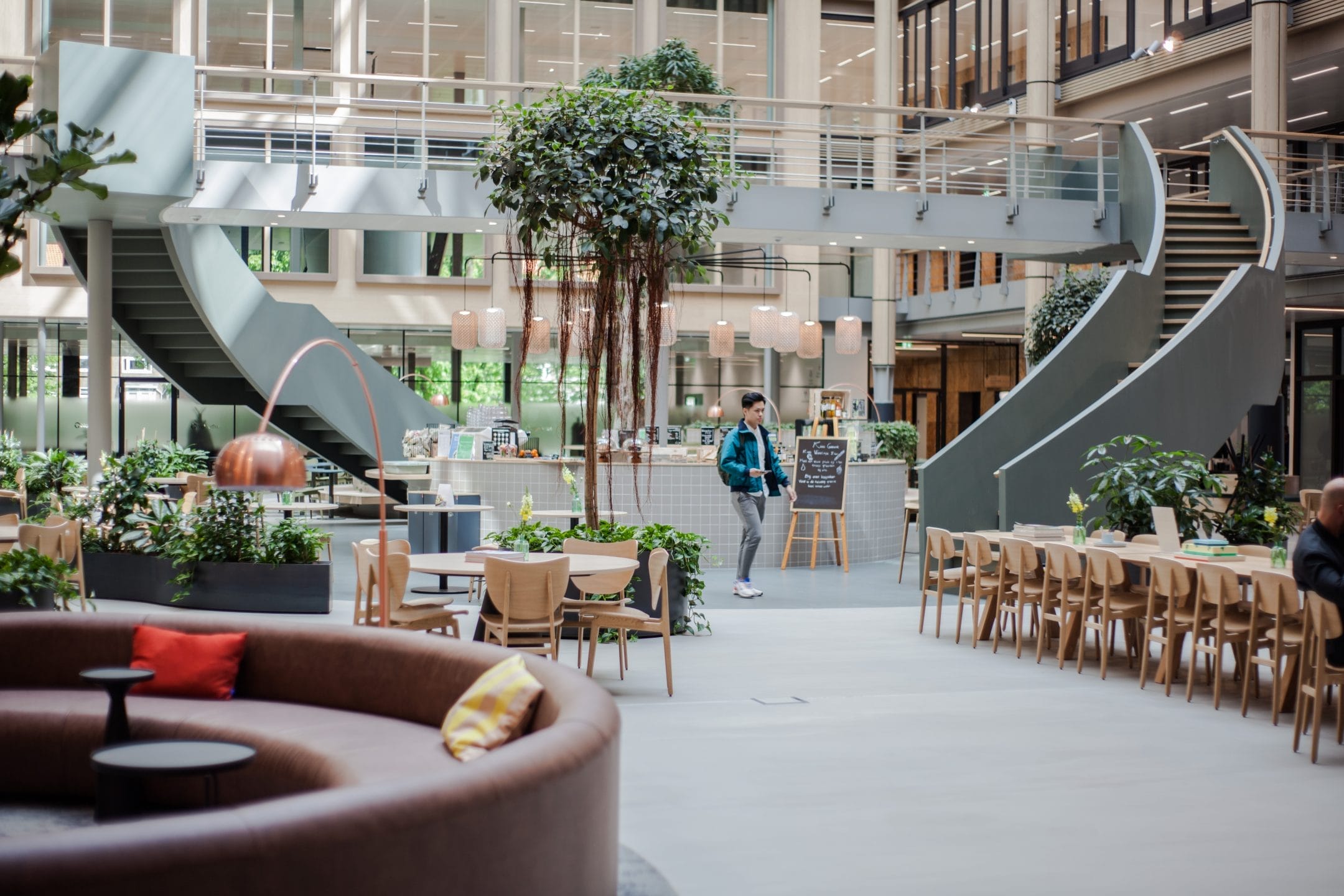 An open-plan office space and coworking spaces with modern architecture featuring a central bar, spiral staircases, ample seating areas, and hanging green plants. Natural light fills the area, enhancing An image from EDGE Workspaces.