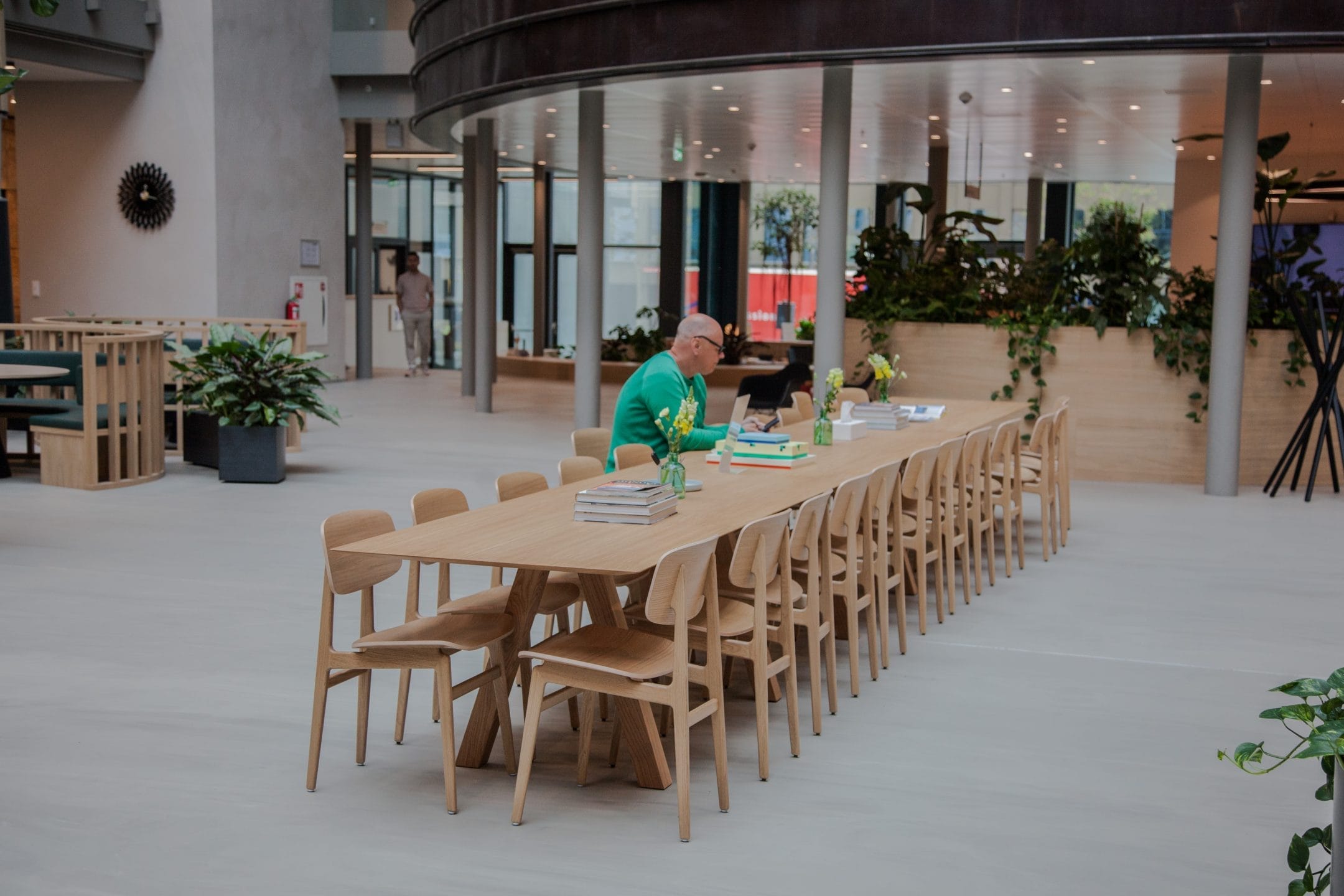 A spacious modern office atrium featuring a long wooden table with matching chairs, ideal for coworking spaces. An older man in a green sweater is reading at the table, surrounded by books and papers, An image from EDGE Workspaces.