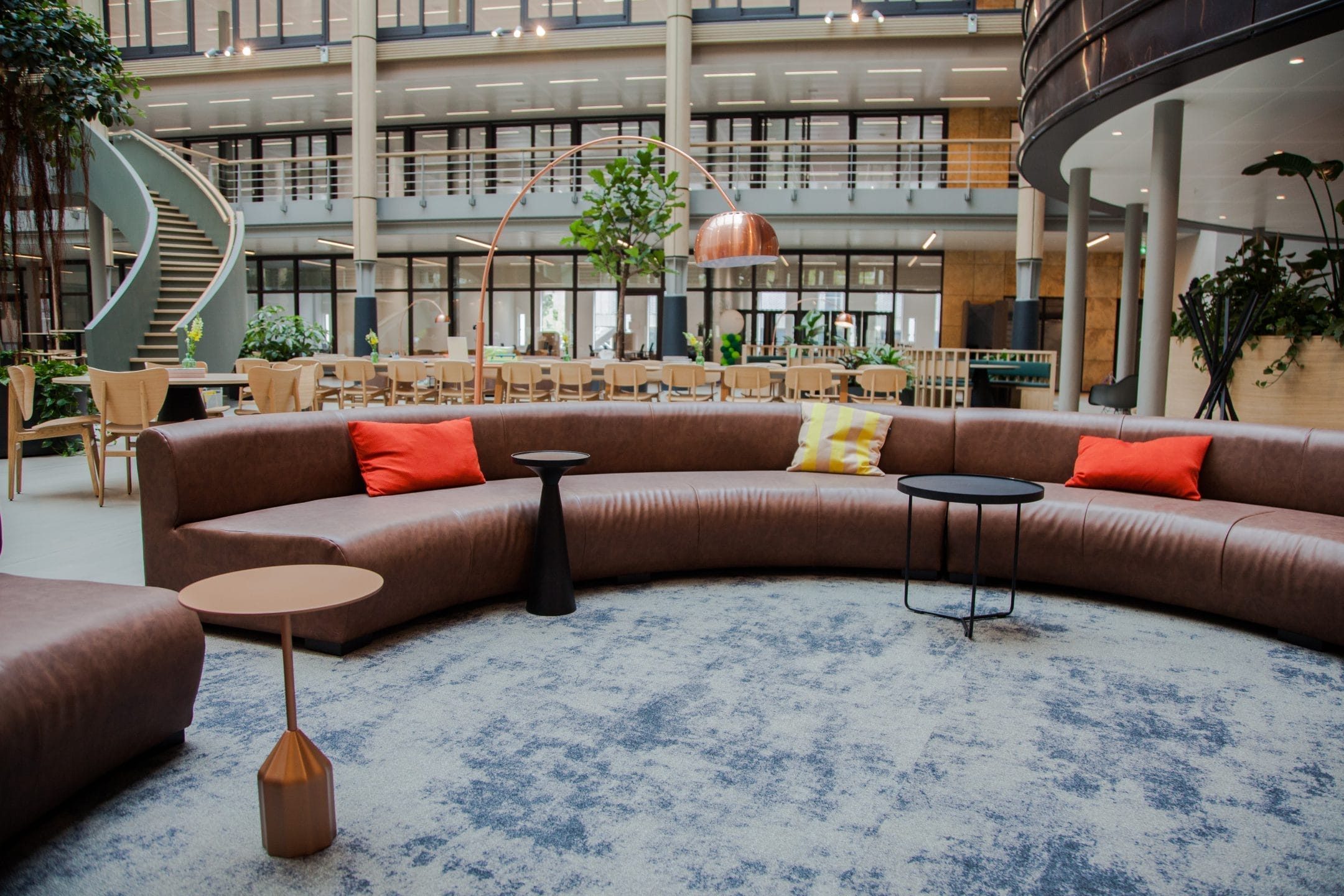 Spacious, modern coworking space with a large, circular brown sofa accented with colorful pillows. Includes round tables, plants, and a spiral staircase in a multi-level building with ample natural light. An image from EDGE Workspaces.