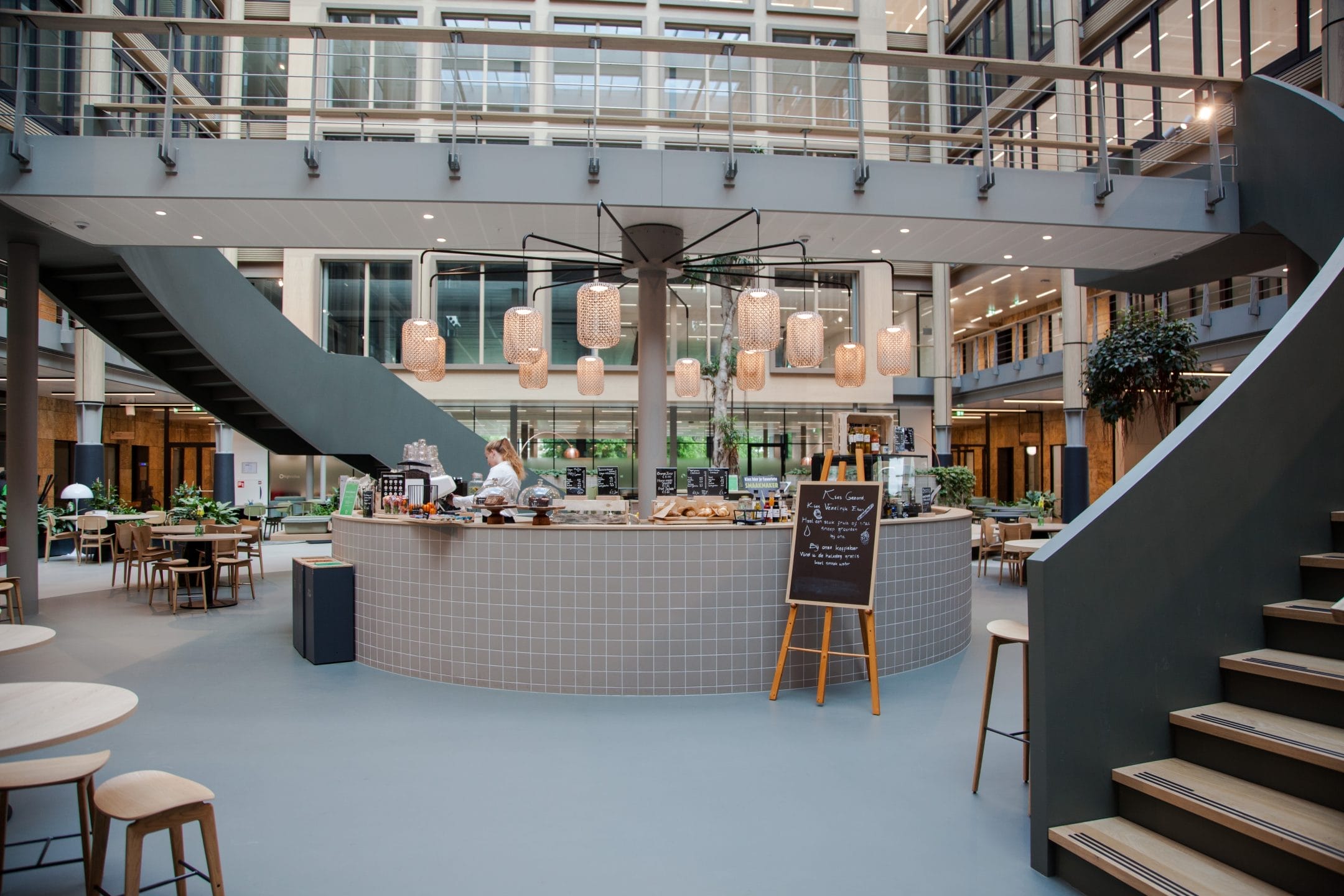 Modern office building cafeteria featuring a circular central serving area with chefs preparing food, surrounded by wooden tables and stylish pendant lights. Staircases on either side lead to innovative coworking spaces on the upper floors An image from EDGE Workspaces.