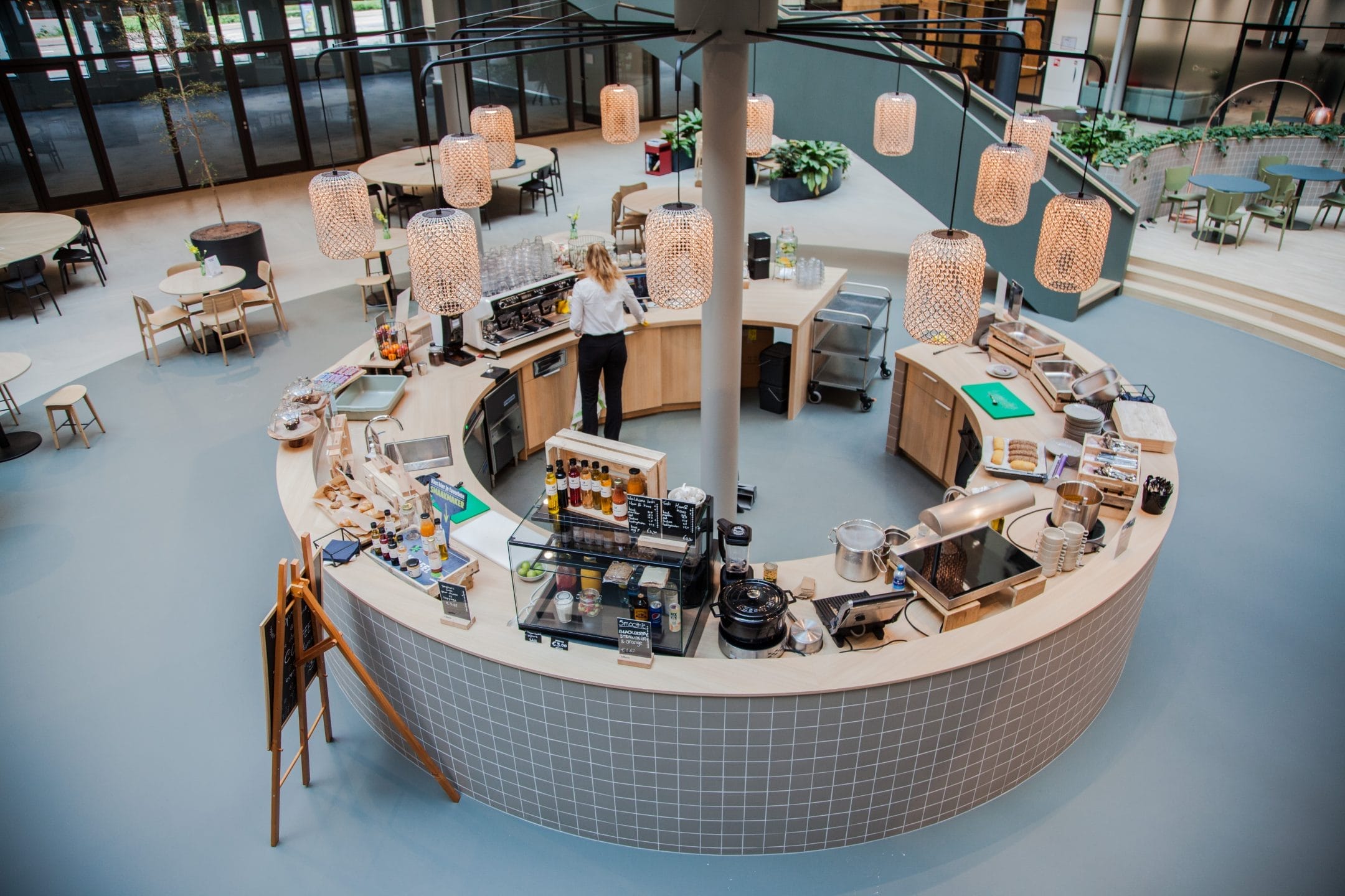 A modern, spacious cafeteria with a circular central serving area and a woman preparing food. The area is surrounded by various dining tables, lush green plant decor, and stylish hanging lamps, creating a welcoming atmosphere An image from EDGE Workspaces.