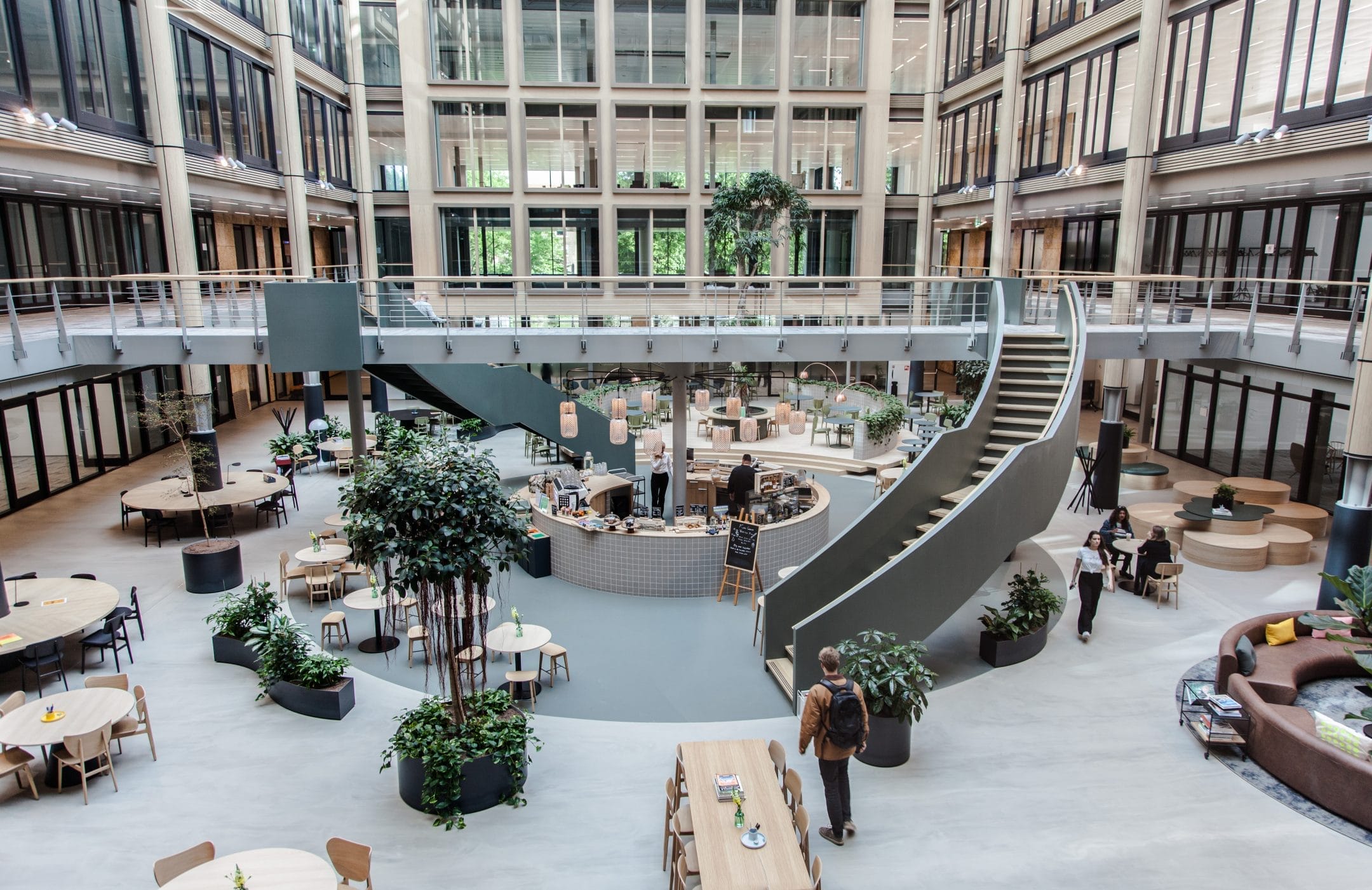 A modern office atrium with a curved staircase leading to upper balconies. The space features a café area with tables and lush plants, surrounded by large glass windows and multiple levels of private office spaces. An image from EDGE Workspaces.