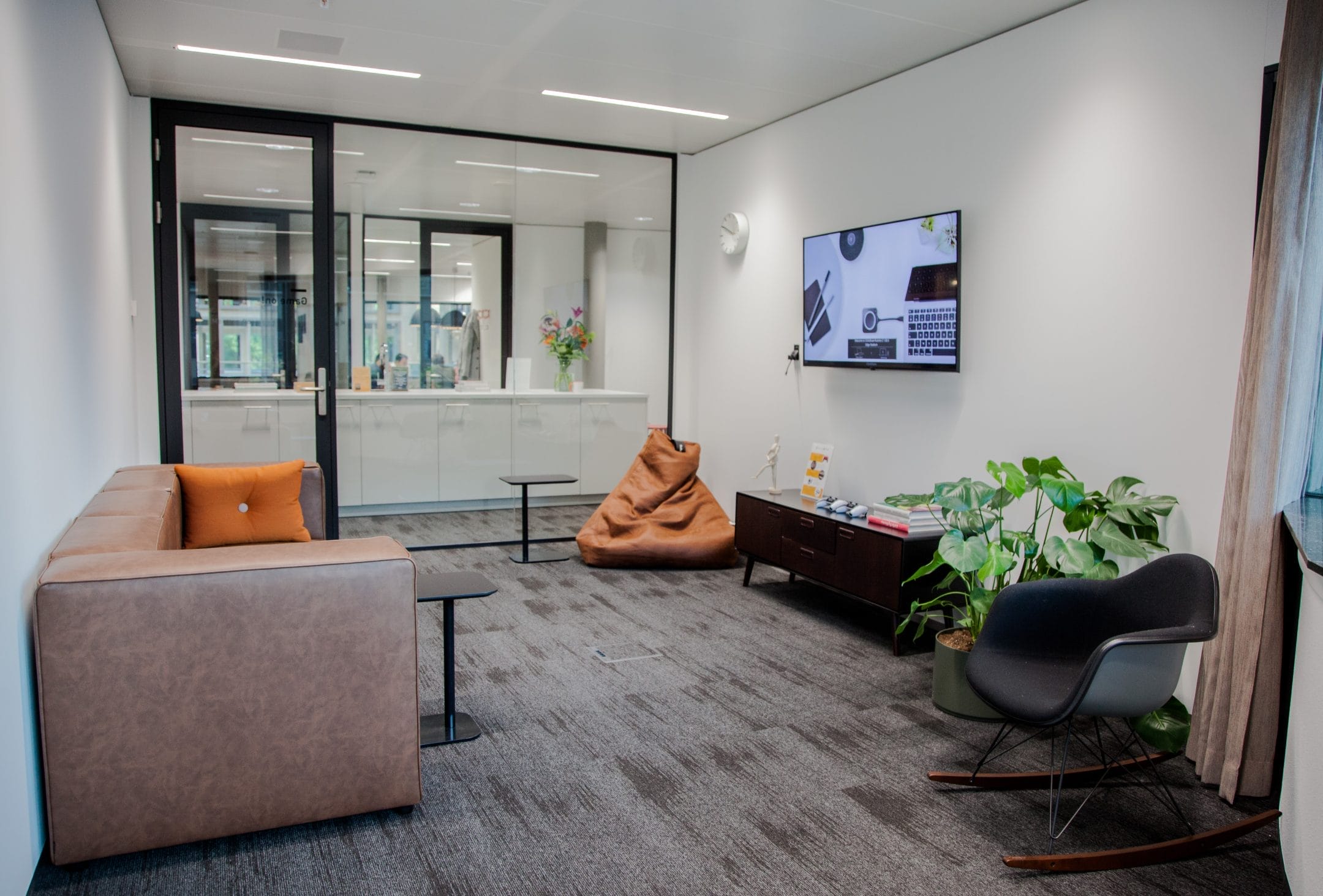 A modern office lounge with a tan sofa, a black rocking chair, a brown bean bag, and a coffee table. There's a large TV on the wall and plants around the room. Glass doors An image from EDGE Workspaces.
