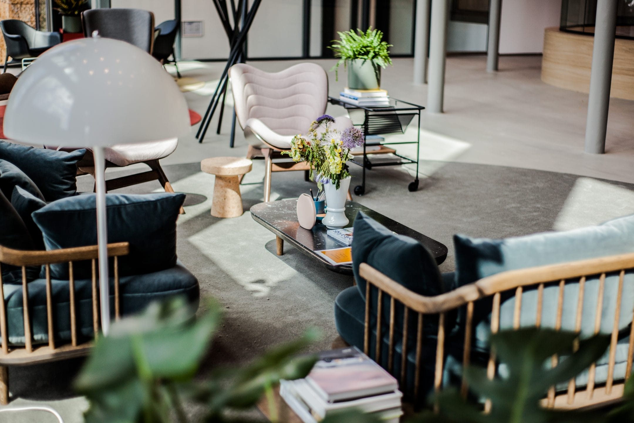 A modern living room featuring a mix of furniture styles including a plush pink chair, blue velvet sofa, and wooden accents. The centerpiece is a coffee table with books and flowers, highlighted by natural lighting— An image from EDGE Workspaces.