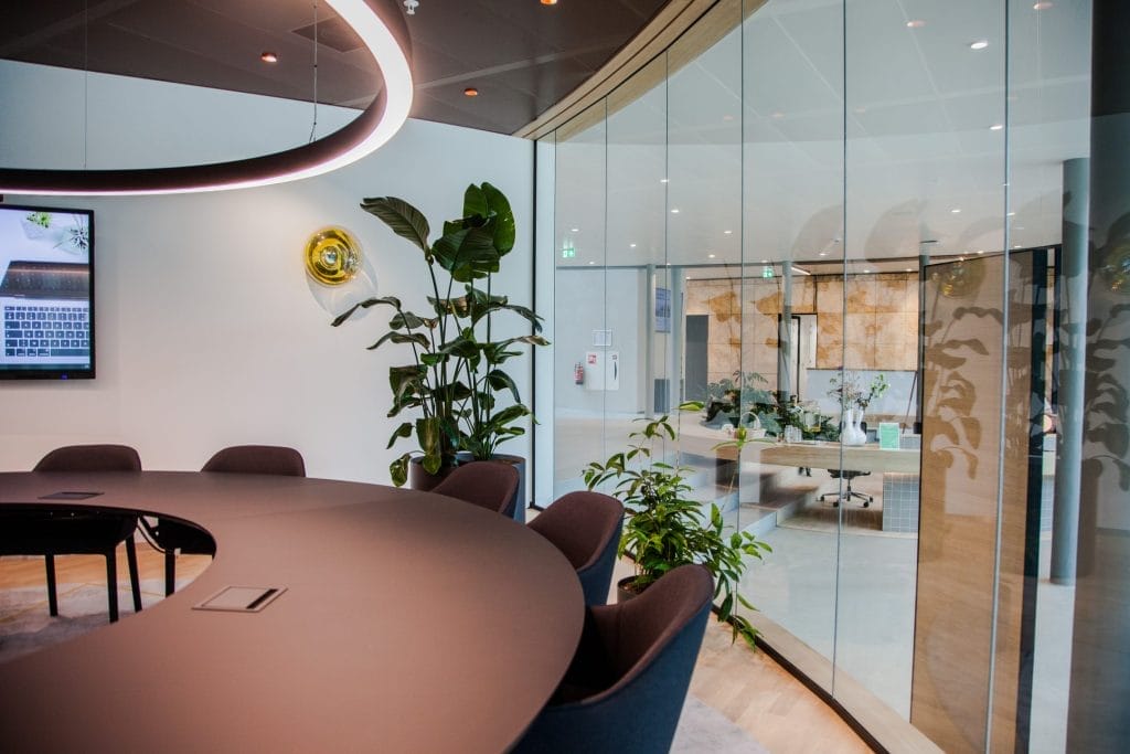 Moderner Besprechungsraum eines Coworking-Büros mit einem großen runden Tisch, Stühlen und einer Topfpflanze. Zu sehen sind Glaswände, ein Fernseher mit Kalenderanzeige und Umgebungsbeleuchtung mit runden Deckenelementen. An image from EDGE Workspaces.