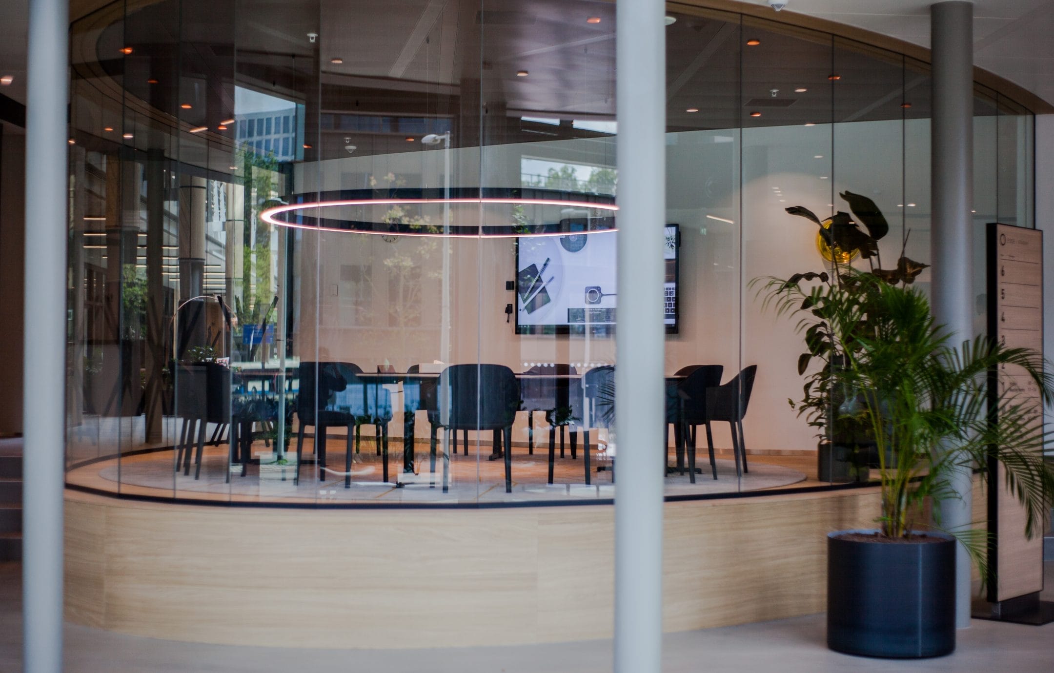 A modern private office space seen through glass walls, featuring a large wooden table with dark chairs, a pink neon circular light overhead, digital screens on the wall, and potted plants adding a touch of An image from EDGE Workspaces.
