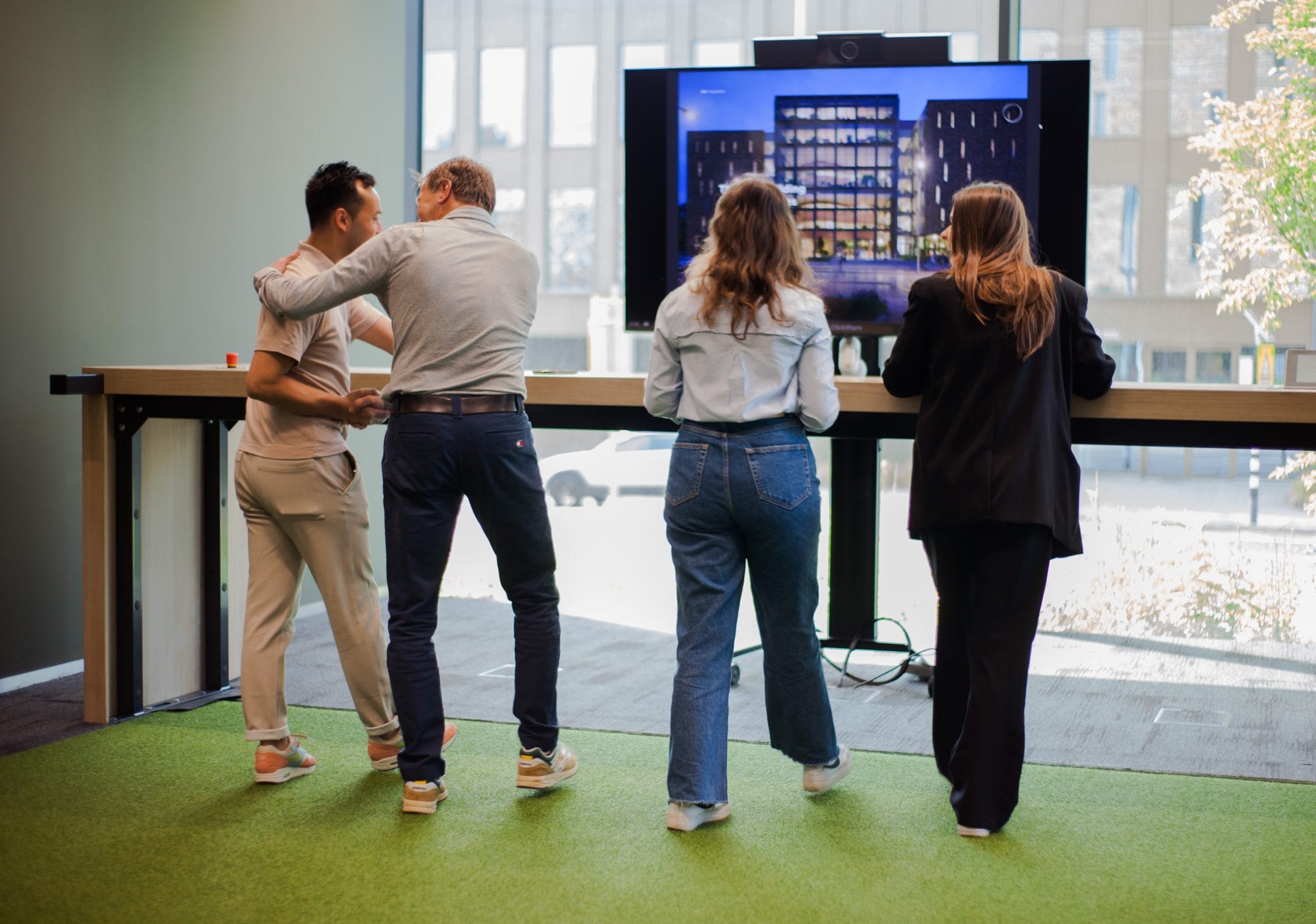Four people stand together in a modern office, watching a large screen displaying a building image. They are engaged in a presentation or discussion, with a focus on the screen, in an innovative coworking environment An image from EDGE Workspaces.