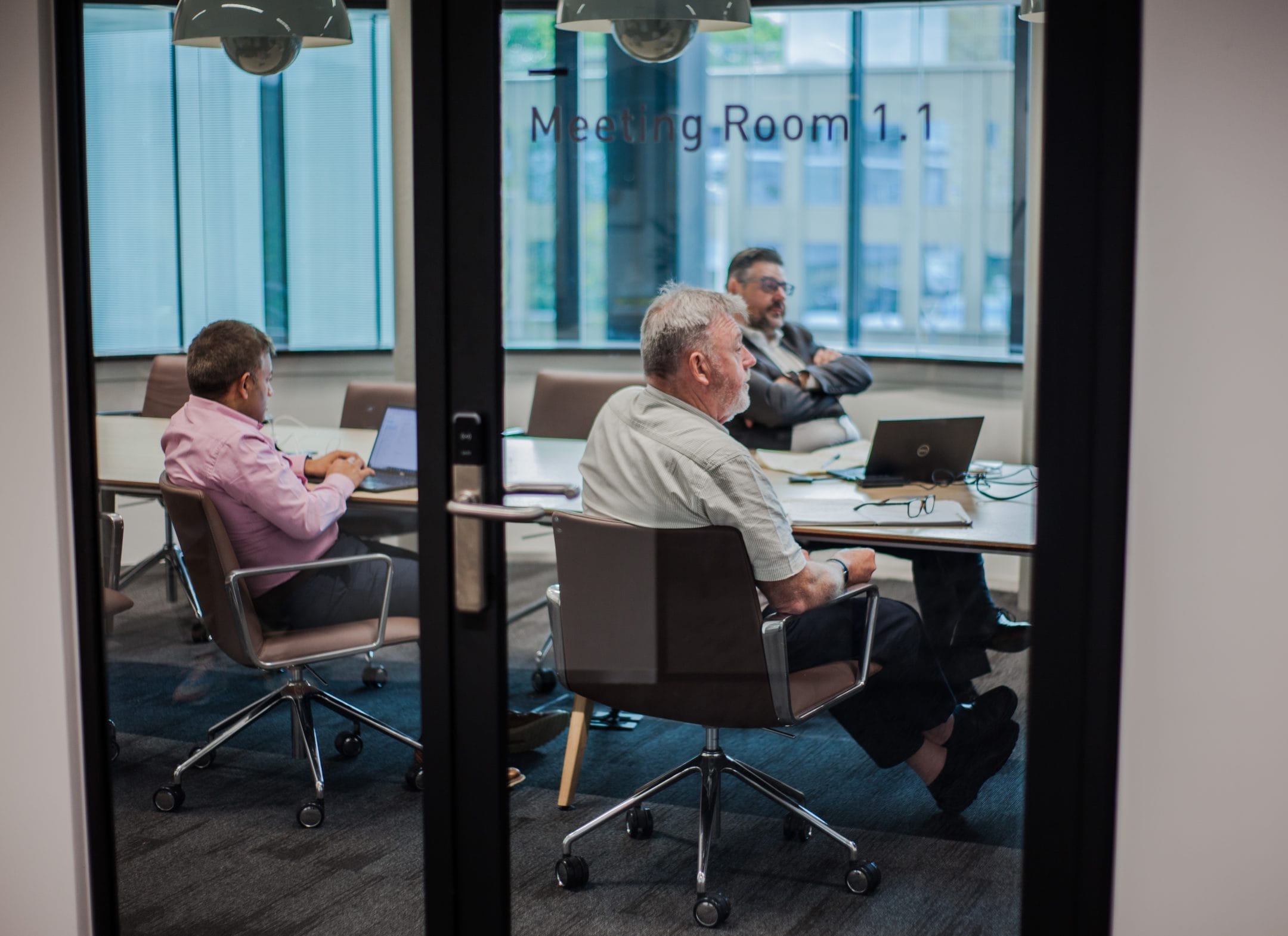 Three men in a modern meeting room, viewed through a glass door marked "meeting room 1.1" in an innovative coworking environment. Two are seated at a table facing a laptop, and An image from EDGE Workspaces.