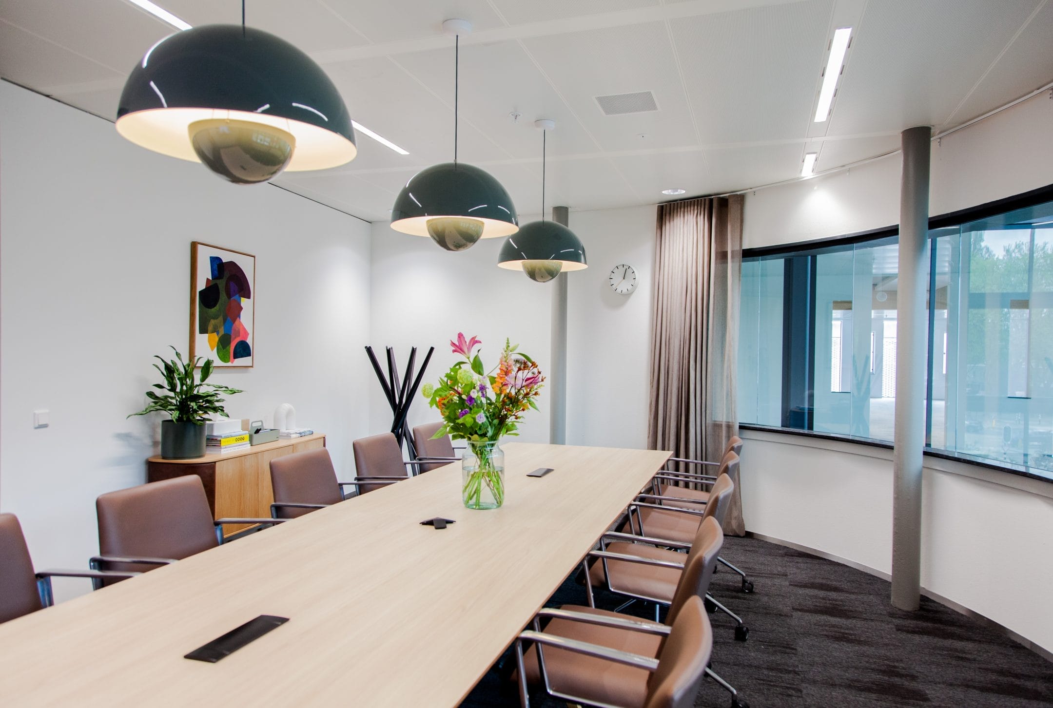 A modern conference room featuring a long table with chairs, three large pendant lights overhead, a colorful abstract painting on one wall, and a vase of fresh flowers on the table. Large windows provide natural light An image from EDGE Workspaces.