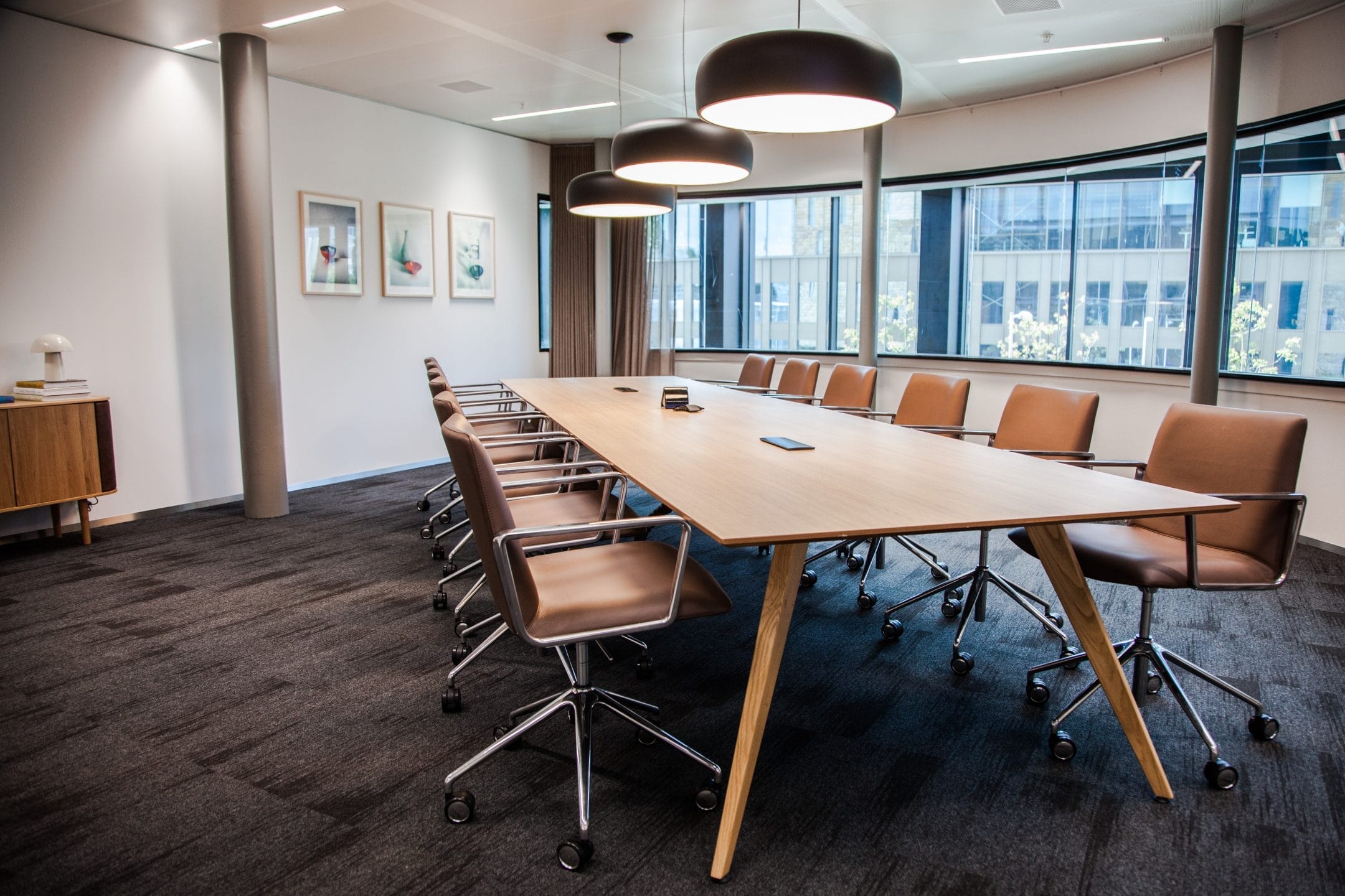 A modern boardroom featuring a long rectangular table with brown leather chairs, surrounded by large windows and adorned with cylindrical overhead lights and framed artwork on the walls, designed for innovative coworking environments. An image from EDGE Workspaces.