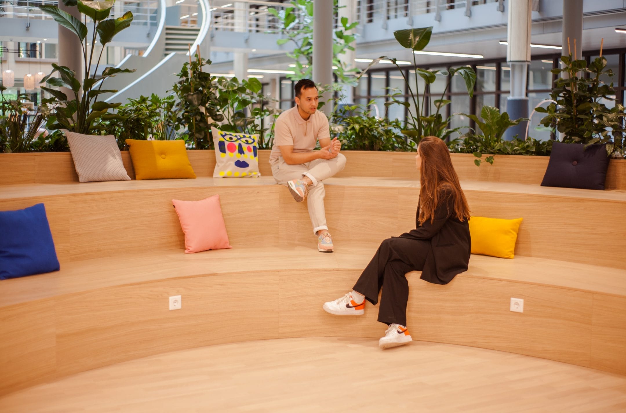 Two people sitting on a wooden amphitheater-style seating area indoors, surrounded by plants, engaged in conversation. The area is well-lit, spacious, and features colorful cushions. This innovative coworking An image from EDGE Workspaces.
