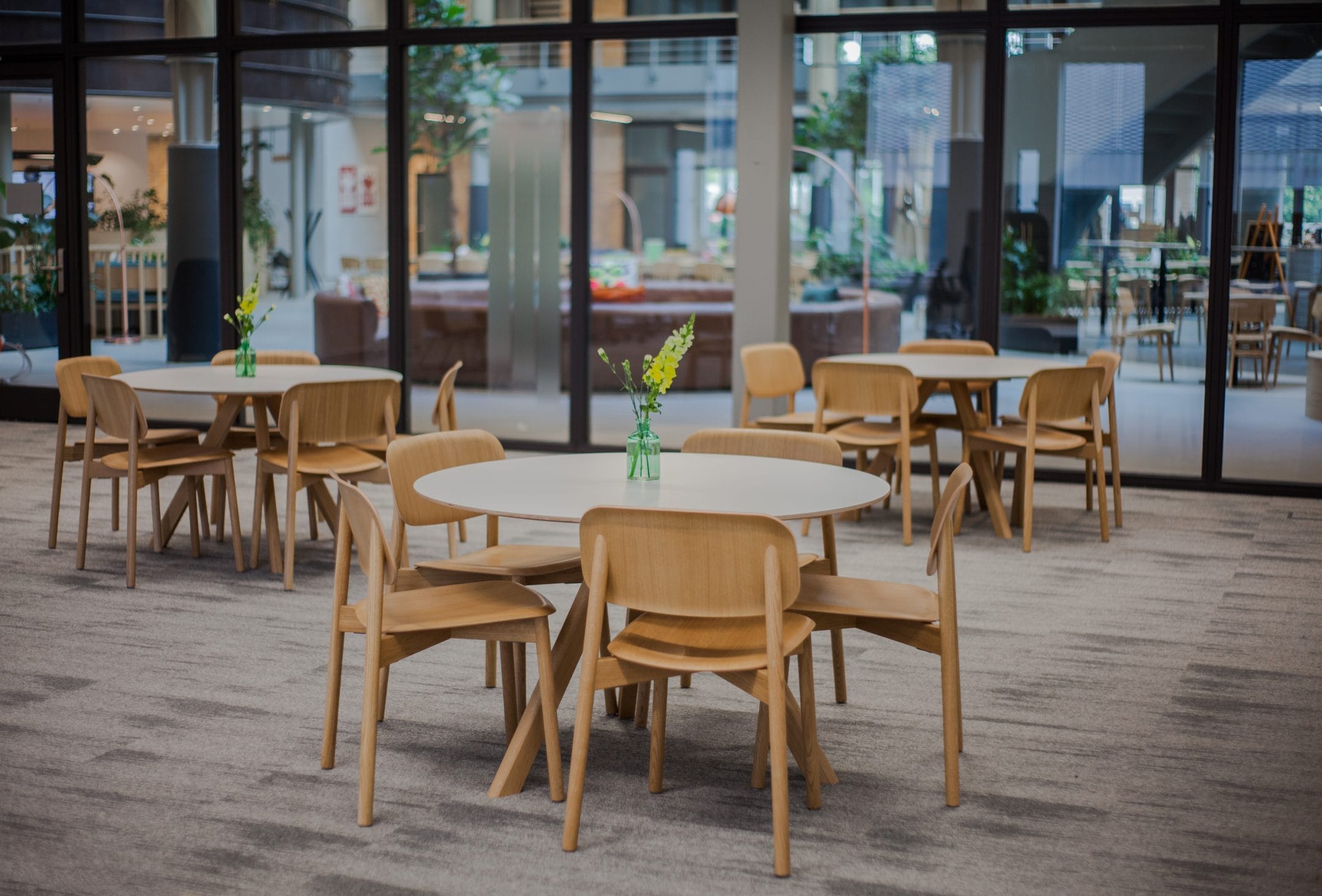 A modern office cafeteria with round tables and wooden chairs serves as a co-working space. Each table has a small vase with green plants. Floor-to-ceiling windows allow natural light to fill the space, An image from EDGE Workspaces.