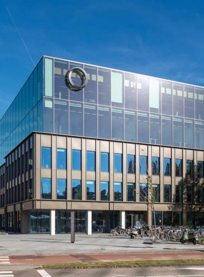 Ein modernes Coworking-Bürogebäude mit reflektierender Glasfassade und einem markanten runden Logo im oberen Bereich. Das Erdgeschoss hat getönte Fenster und sichtbare Säulen, draußen parken Fahrräder unter einem klaren blauen An image from EDGE Workspaces.