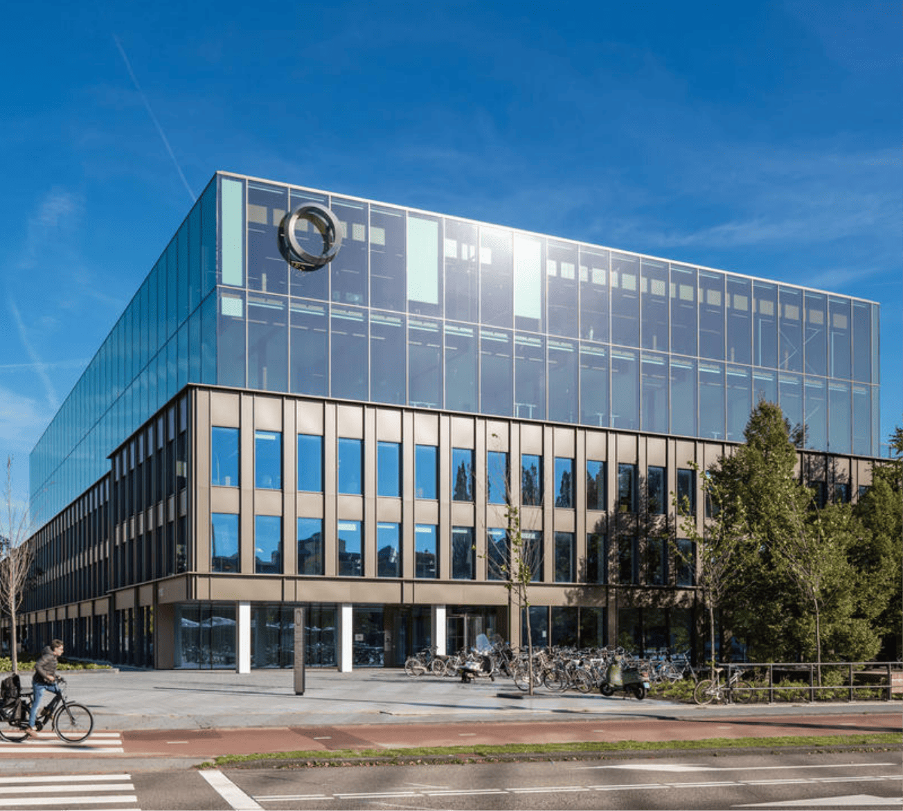 Ein modernes, mehrstöckiges Bürogebäude mit umfangreichen Glasverkleidungen und einem prominenten Logo oben in der Mitte bietet Mitgliedschaften für dedizierte Arbeitsbereiche und private Büros. Radfahrer und Fußgänger gehen im Vordergrund neben geparkten An image from EDGE Workspaces.