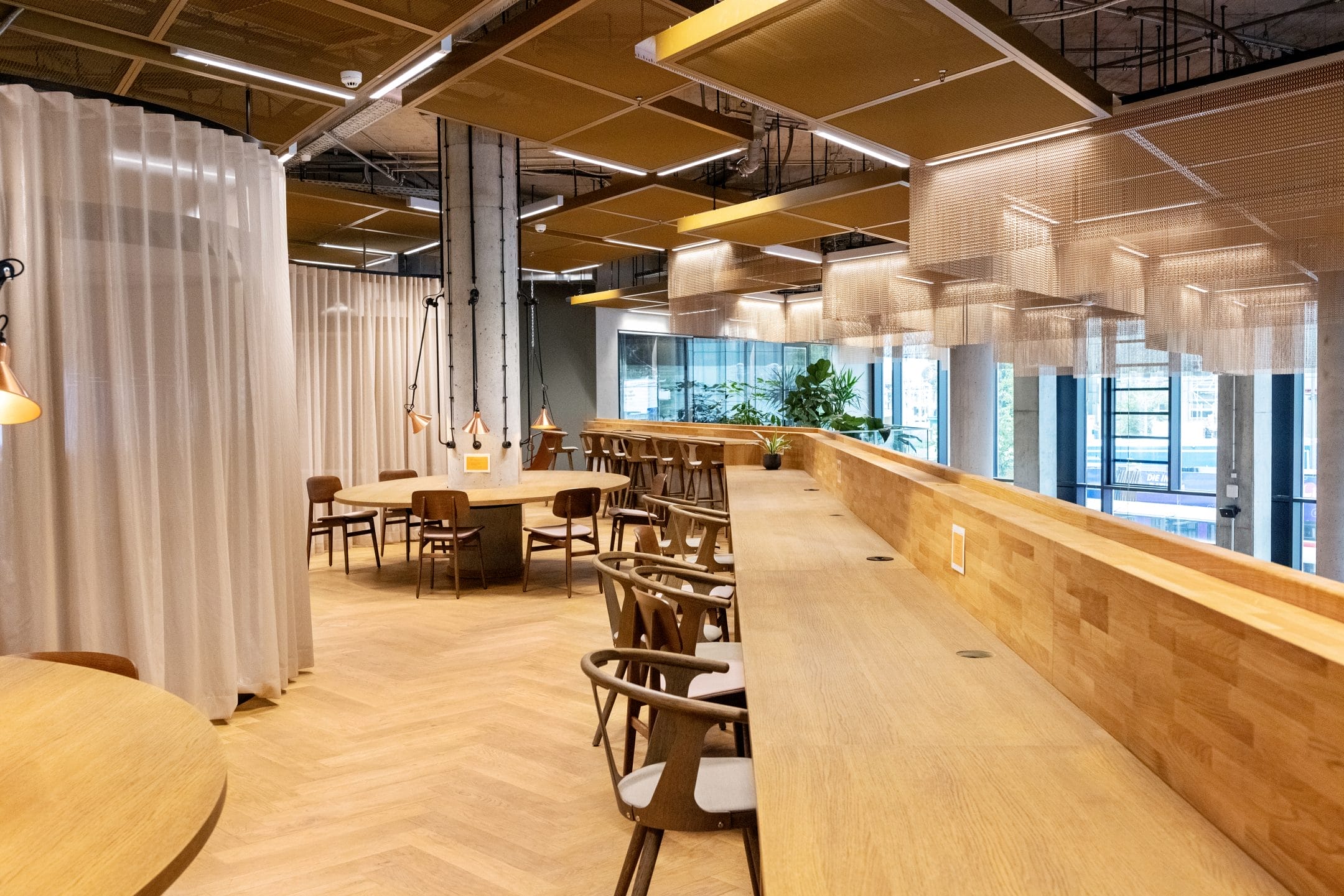 A modern, well-lit coworking space featuring wooden tables and chairs, a long bar with stools, and ceiling-hanging lamps. Sheer curtains separate areas, with plants visible through a large window An image from EDGE Workspaces.