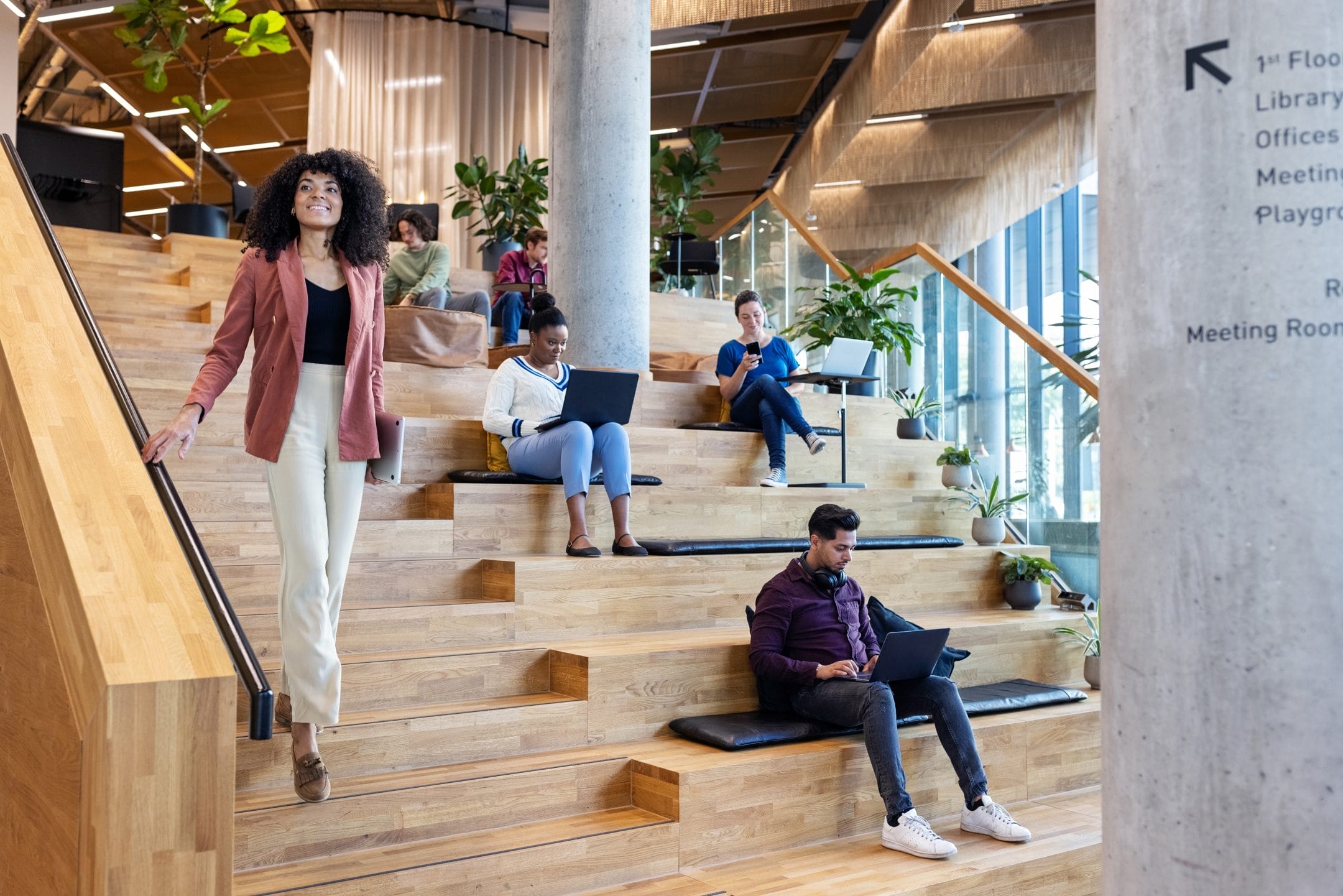 A diverse group of people in a modern office stairway space, using it as an innovative coworking environment for different activities like walking, sitting, and working on laptops, with directional signs overhead. An image from EDGE Workspaces.