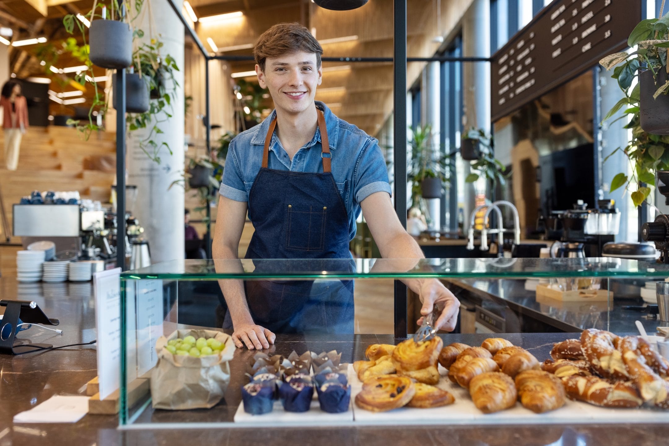 A cheerful young man in a denim apron stands behind a bakery counter, offering freshly baked goods such as croissants and muffins in an innovative coworking environment, with hanging plants and a blurred An image from EDGE Workspaces.