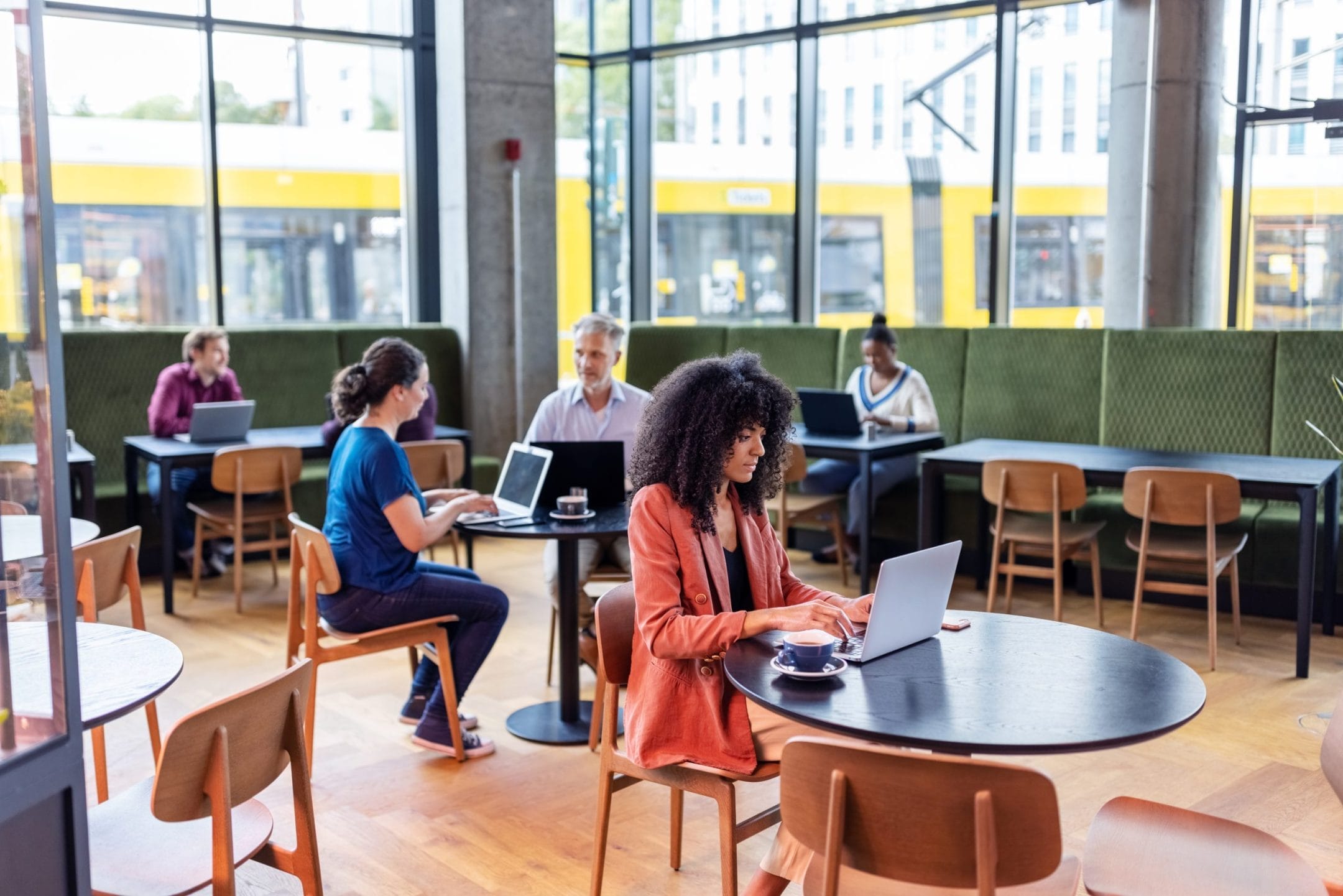 A young woman with curly hair works on a laptop in a brightly lit modern coworking space with large windows. Other patrons are seated around her, also engaged in work on their laptops, in a relaxed An image from EDGE Workspaces.