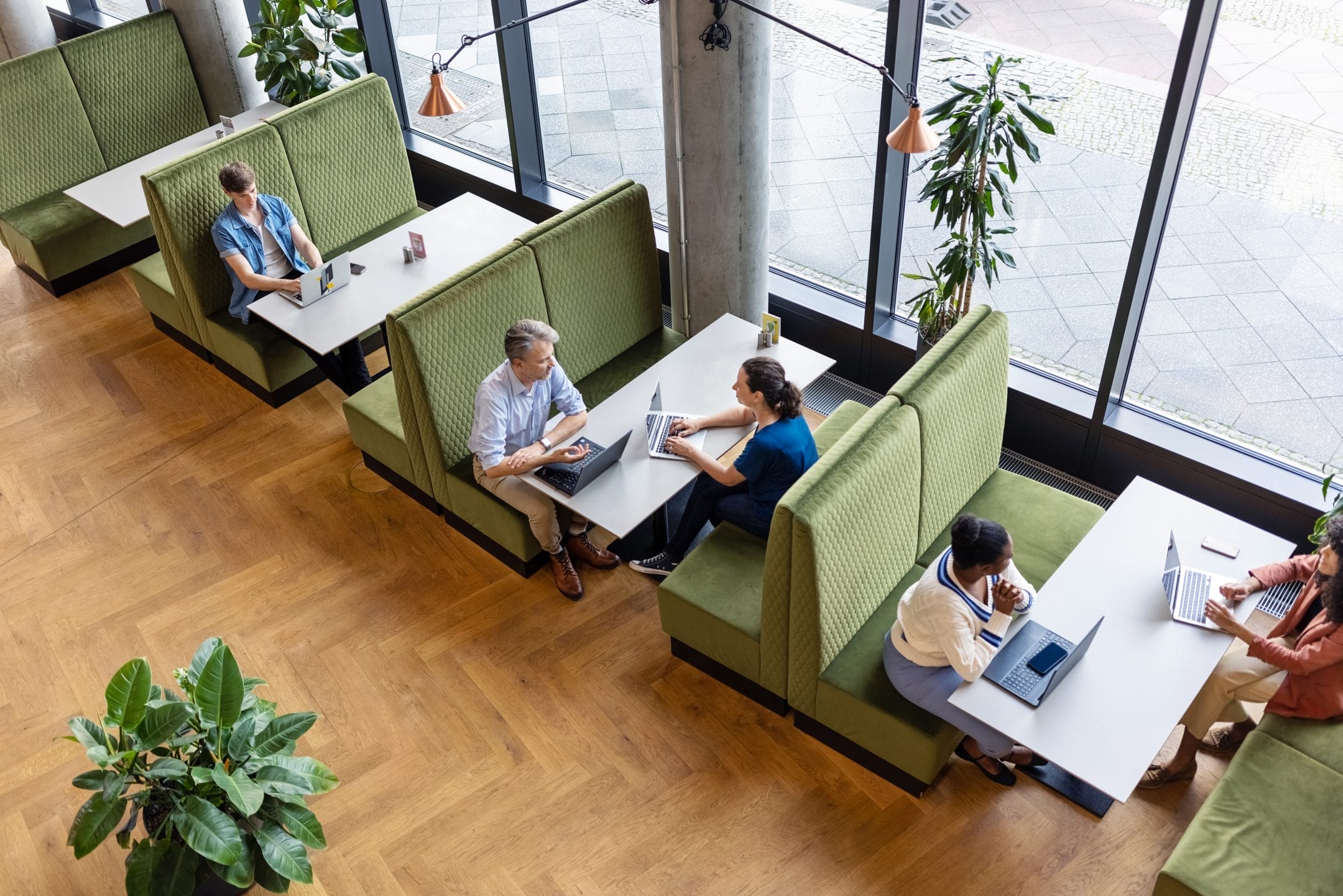Overhead view of an innovative coworking environment with green booths where individuals, both alone and in pairs, work on laptops. The space is bright, surrounded by large windows and decorated with plants. An image from EDGE Workspaces.