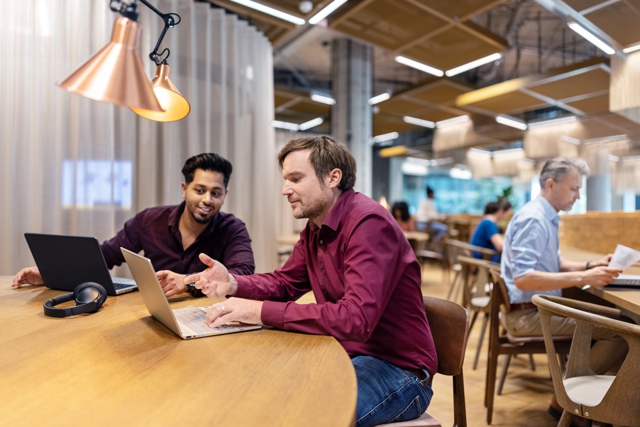 Two men, one South Asian and one Caucasian, collaborate on a laptop at a co-working space with modern decor, offering flexible workspace solutions. A third man works quietly in the background. An image from EDGE Workspaces.