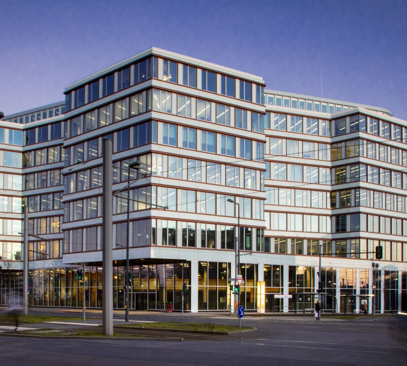 Moderner mehrstöckiger Coworking-Space mit Glasfassade und strukturierten weißen Rahmen, gelegen an einer Straßenecke unter einem klaren blauen Abendhimmel mit sanfter Beleuchtung. An image from EDGE Workspaces.