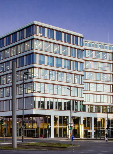 A modern multi-story office building with reflective glass windows and white framing under a clear blue sky. The building's ground floor is visible with coworking spaces, accessible entrances, and parking spaces at the front An image from EDGE Workspaces.