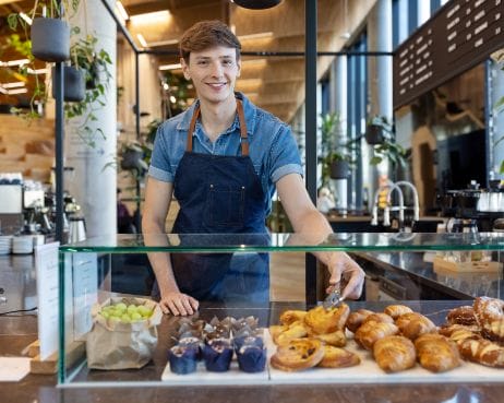 Ein junger männlicher Bäckereiangestellter, der eine Jeansschürze trägt, lächelt herzlich, während er hinter einer Auslage mit einer Auswahl an Gebäck wie Croissants und Schokoladenleckereien in einer modernen Bäckereikuh steht An image from EDGE Workspaces.