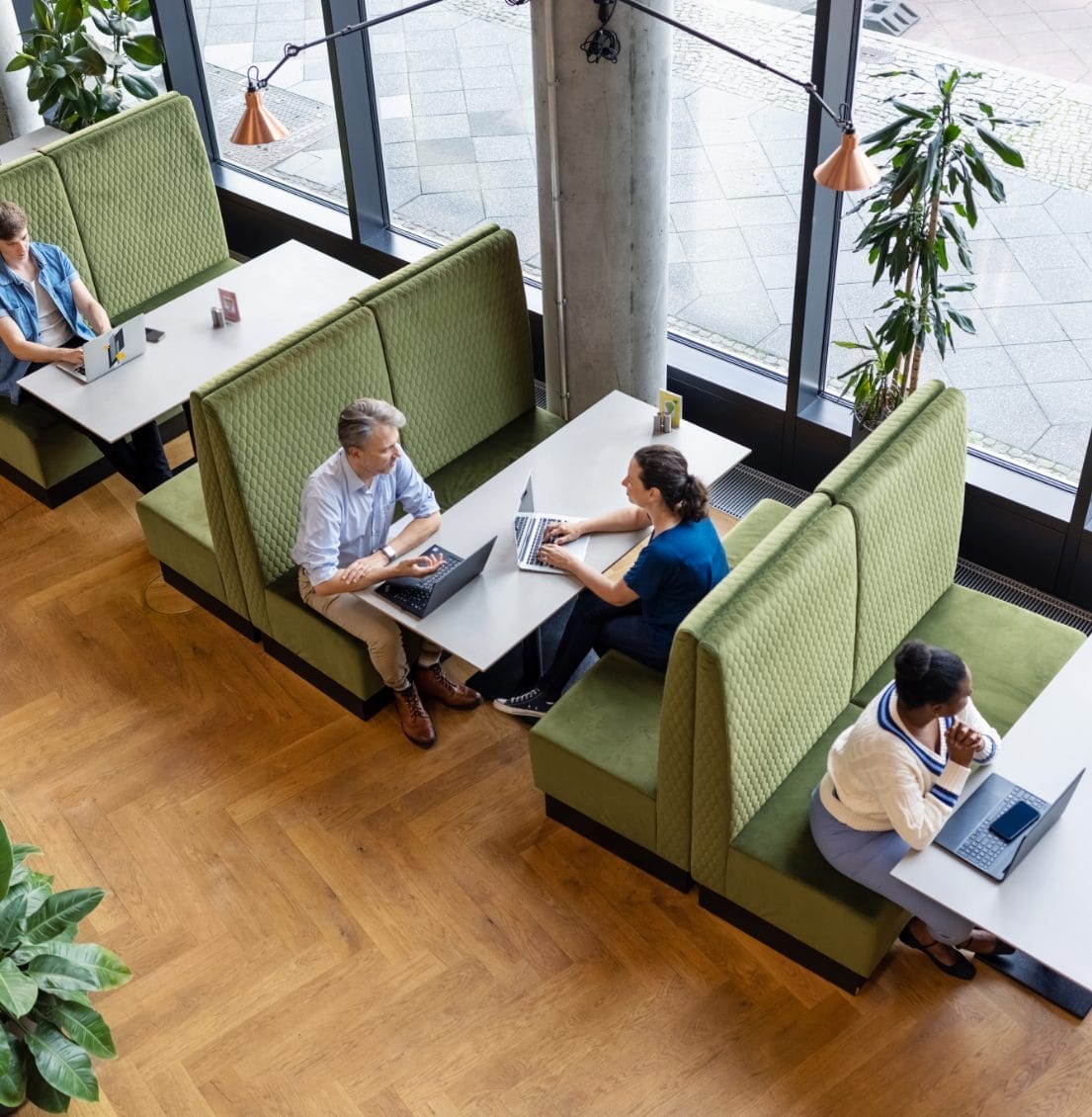 Luftaufnahme eines modernen Coworking Space mit vier Personen, die an getrennten Arbeitsplätzen arbeiten. Zwei sind an einem Tisch in eine Diskussion vertieft, während die anderen unabhängig voneinander an Laptops arbeiten. Der Raum verfügt über große Fenster An image from EDGE Workspaces.