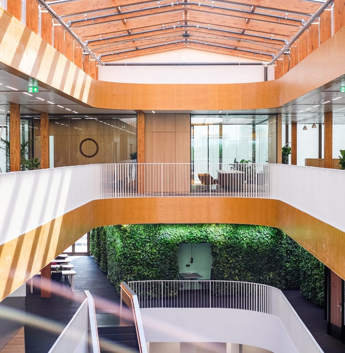 Modern office building interior with curved white walkways, wooden paneling, and a lush green plant wall. The design features abundant natural light, minimalist decor, and innovative coworking environments. An image from EDGE Workspaces.