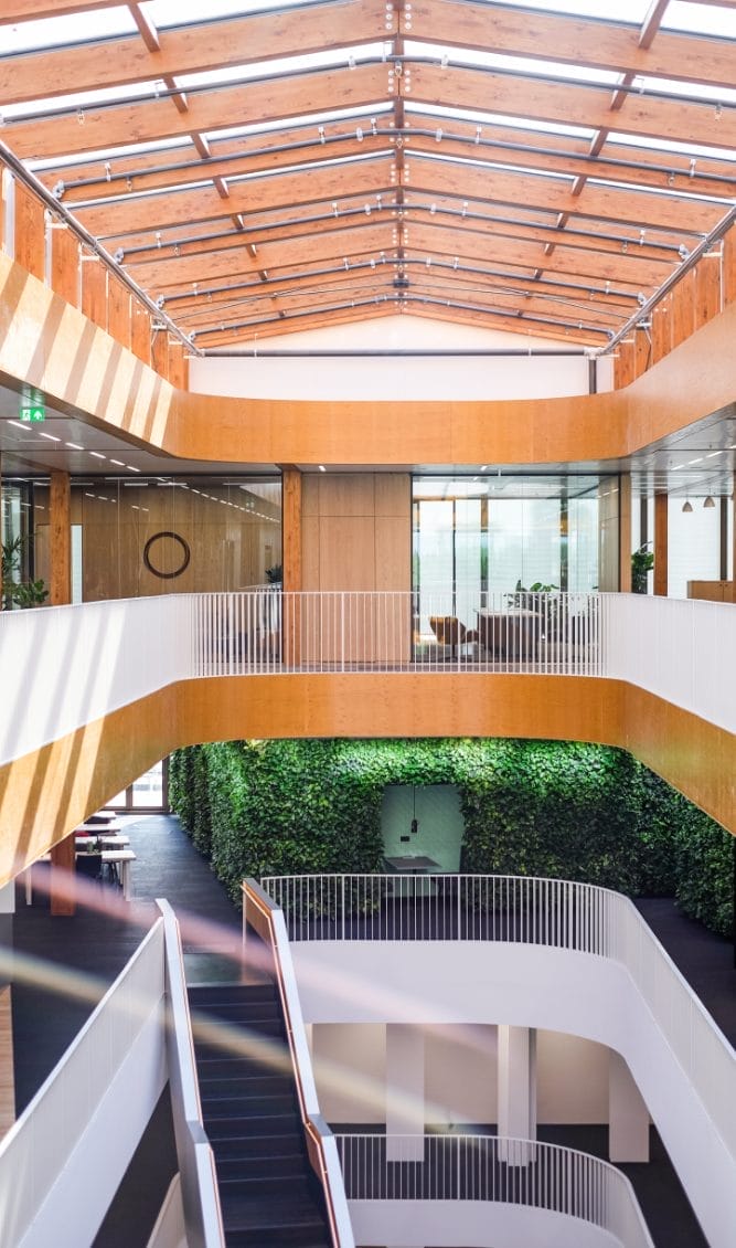 Modernes Bürogebäude mit vertikalem Design, einem zentralen Atrium mit Glasdecke, Holzbalkonen, grünen Wänden und weißen Treppen, die die verschiedenen Stockwerke verbinden. Es umfasst spezielle Arbeits An image from EDGE Workspaces.