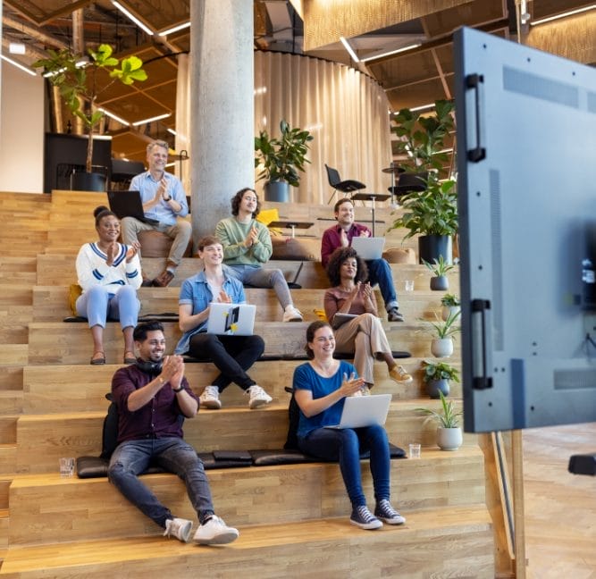 A diverse group of seven people sitting on wooden bleacher-style seating in a coworking space, watching and clapping at a presentation shown on a screen, visible from the back. The room has green An image from EDGE Workspaces.