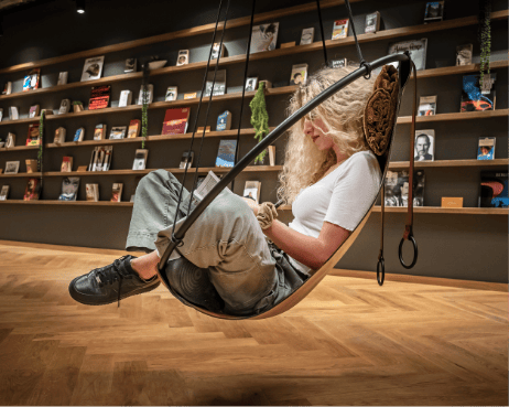 Eine Frau mit lockigem Haar sitzt auf einer stilvollen, geschwungenen Schaukel in einem modernen Buchladen, der gleichzeitig als Coworking-Space dient, umgeben von Wandregalen voller Bücher. Sie wirkt entspannt und blickt An image from EDGE Workspaces.