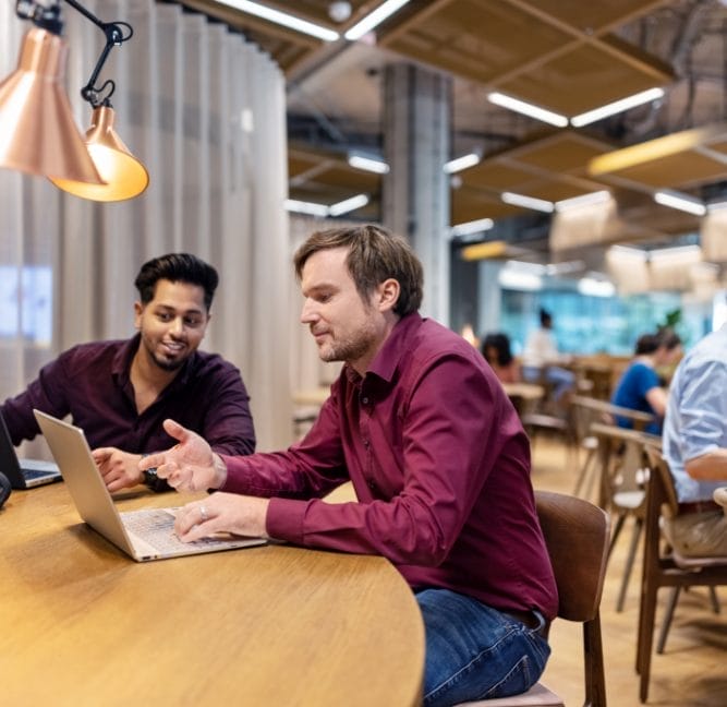 Two men sit at a table in an innovative coworking environment, working on a laptop. One man, who appears engaged, is pointing at the screen while explaining something to the other, who is listening An image from EDGE Workspaces.