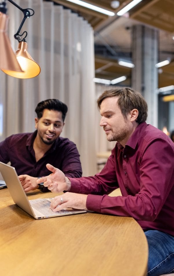 Two men, one with dark hair and one with light hair, are sitting at a wooden table in a well-lit private office. They are engaged in a discussion with a laptop open in front of An image from EDGE Workspaces.