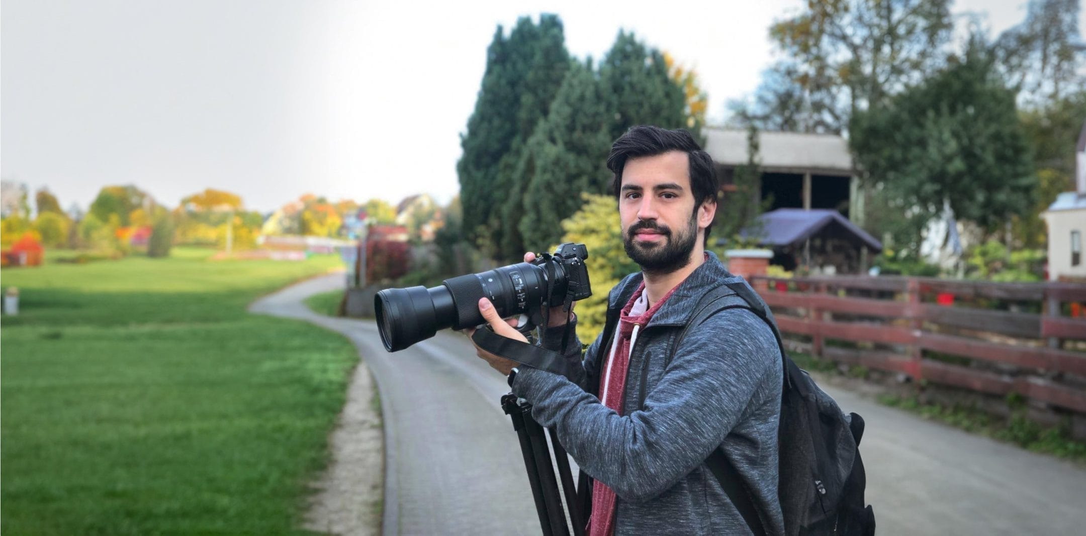 Ein bärtiger Mann mit einer Kamera mit großem Objektiv steht auf einem Weg in der Nähe eines Bauernhofs mit grünen Feldern und Veranstaltungsräumen im Hintergrund. Er trägt einen Rucksack und ein kariertes Hemd unter An image from EDGE Workspaces.