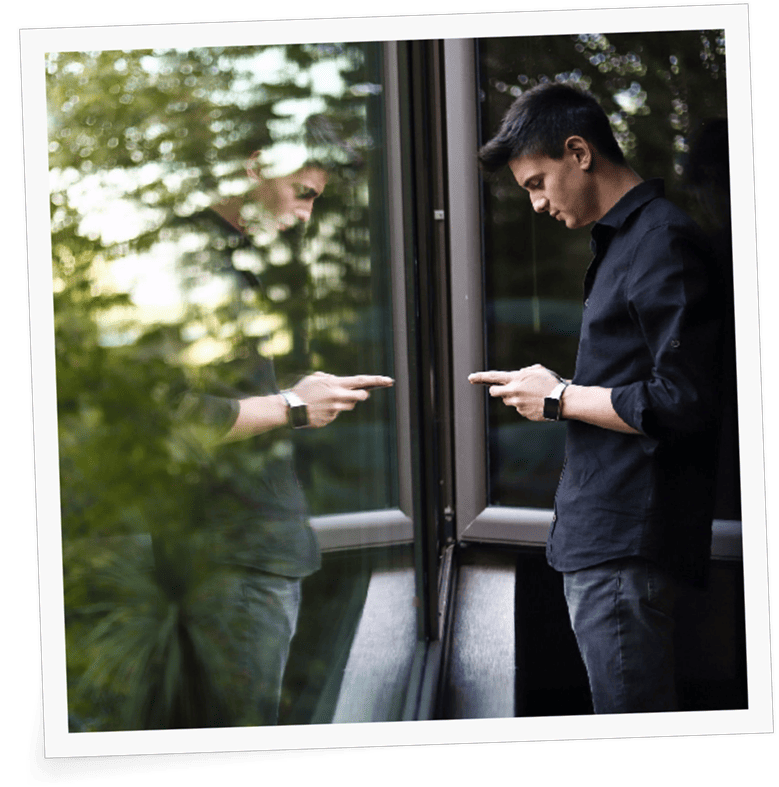 Ein junger Mann in dunklem Hemd steht an einem Fenster in einem Coworking Space und berührt das Glas mit dem Finger, während sein Spiegelbild dies widerspiegelt. Im Hintergrund sind verschwommenes Grün und Fensterrahmen zu sehen. An image from EDGE Workspaces.